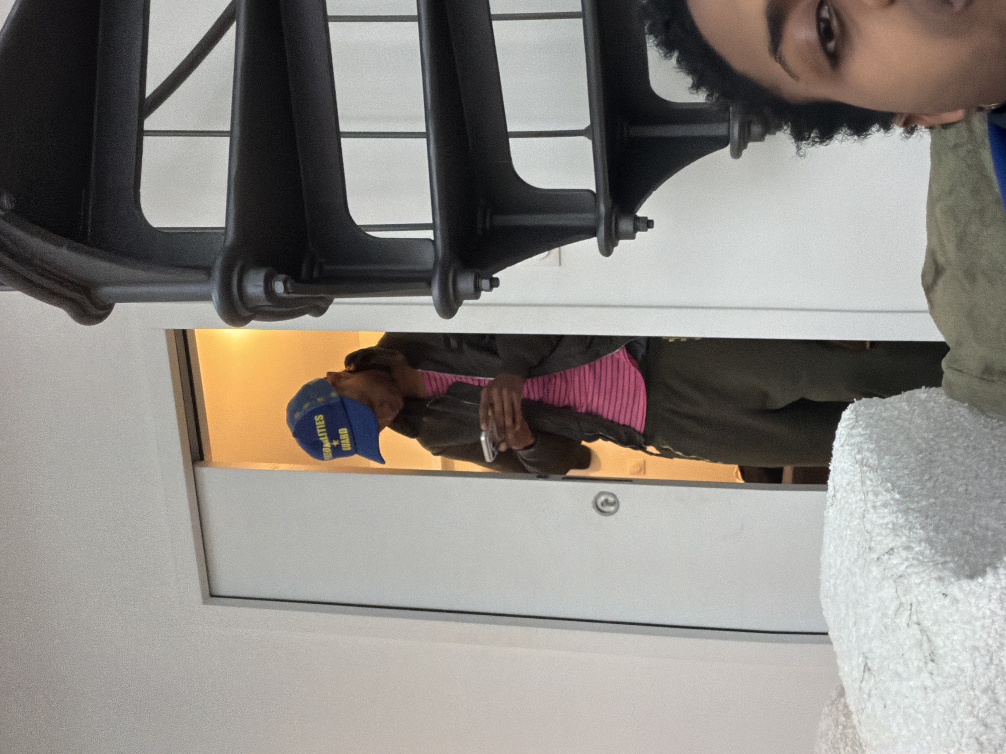 A person wearing a blue "PERSONALITIES GUARD" hat, a pink striped shirt, and an olive green jacket is standing in a doorway. The person is holding a phone in their hands. The setting appears to be indoors, possibly a residential or office space with a white door and a metallic radiator in the foreground. Another person's face is visible in the top right corner. The location is unconfirmed Saint-Denis, France.