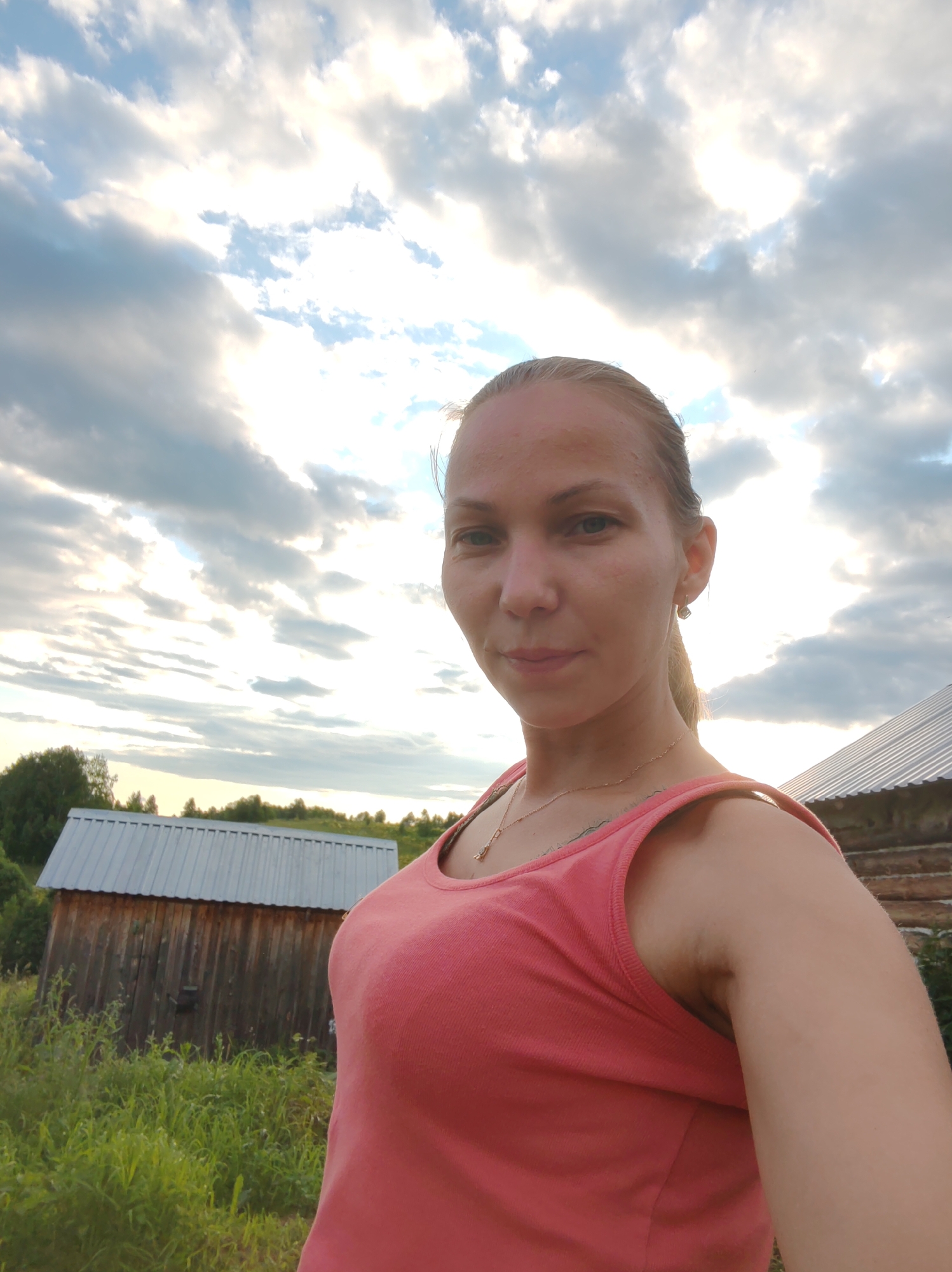 In a medium shot, a Caucasian woman with blonde hair tied back in a ponytail is looking at the camera. She is wearing a coral pink tank top and a delicate gold necklace. Her right shoulder and arm are visible, as is a hint of a tattoo on her upper chest. She has light skin and appears to be in her late 20s or early 30s. She is standing outside, with a wooden outbuilding to her left and a section of a rustic wooden building to her right. The sky above is filled with dramatic, grey and white clouds, suggesting either an overcast day or late afternoon. The ground is covered in lush green grass and vegetation. The location is identified as the village of Chukhlem, Russia.