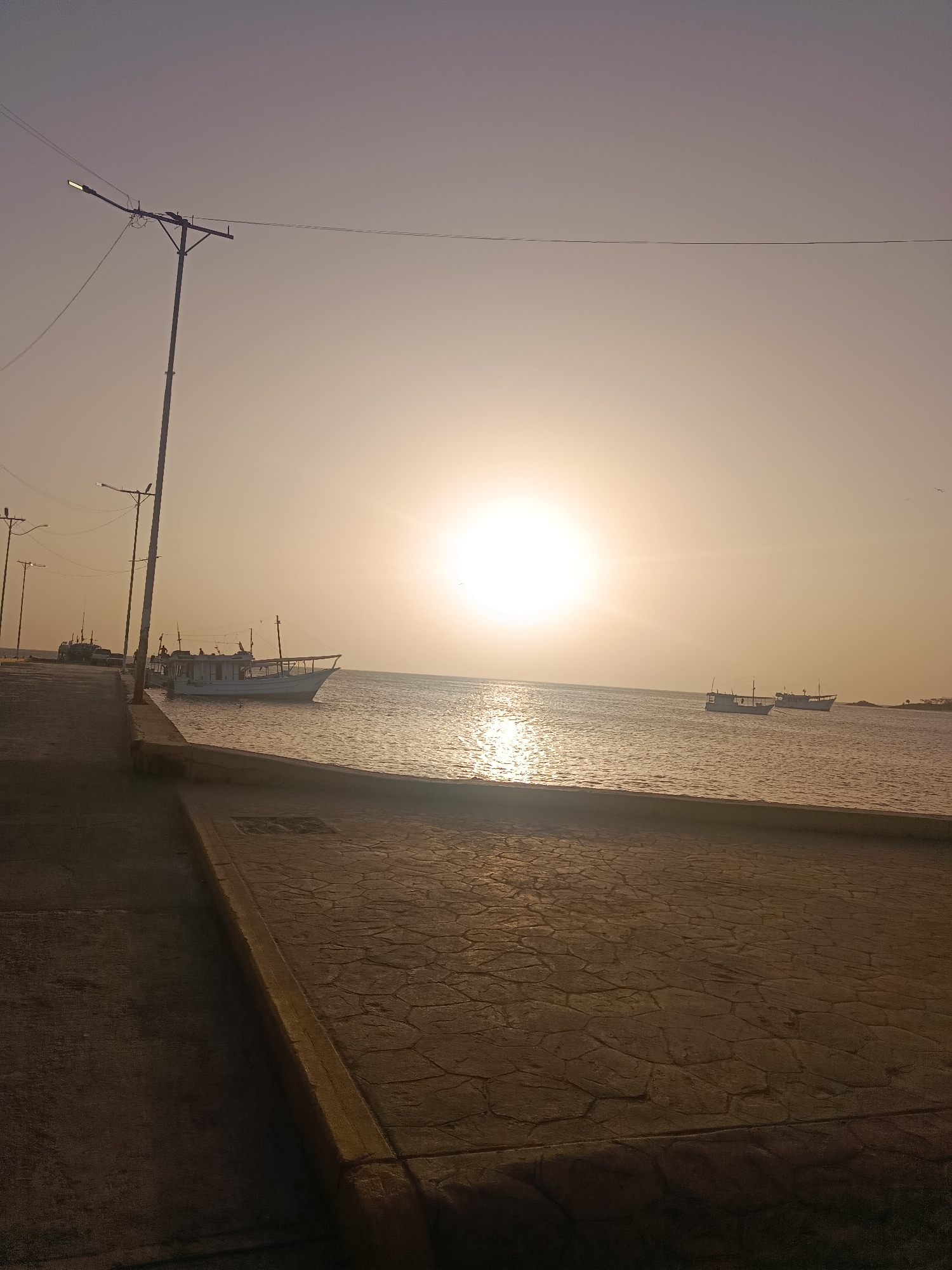 This image depicts a view of the sea from a paved area, likely a promenade or harbor in Juan Griego, Venezuela. The sun is low on the horizon, creating a golden hue in the sky and reflecting on the water. Several fishing boats are visible in the water. There are utility poles lining the paved area. The paving consists of concrete and a patterned surface.