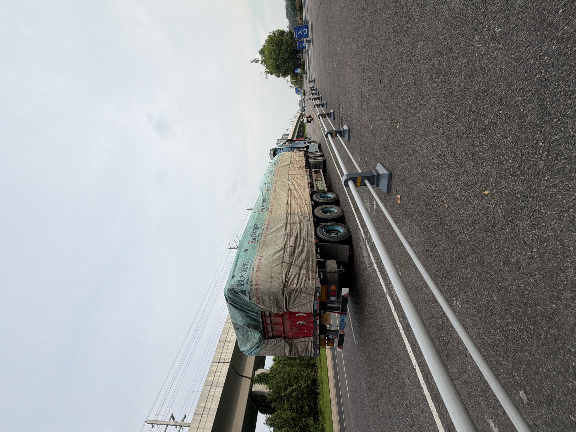 A heavy-duty truck with a tarp-covered load is traveling on a highway in Jiangjin, China. The truck, which appears to be a semi-trailer, is covered with a large, greenish-brown tarp secured with ropes. Chinese characters are visible on the tarp. The truck is in the right lane of a paved highway. In the background, an elevated concrete structure, possibly a bridge or overpass, is visible with power lines running alongside it. Trees and vegetation line the side of the highway. Further in the distance, there is a cell tower and a sign with Chinese characters and a parking symbol, indicating a nearby entrance. The sky is overcast and appears to be daytime. A few other vehicles and a motorcycle are also visible on the highway in the distance. The road is lined with reflective barriers.