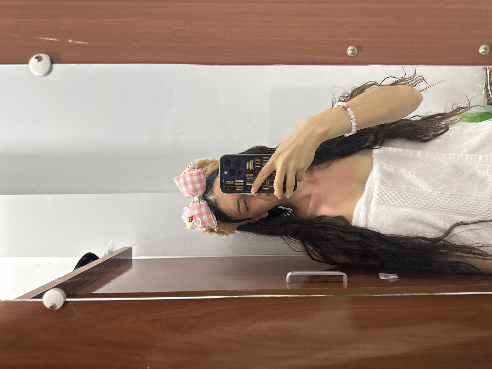 A mirror selfie shows a person with long brown hair wearing a pink and white checkered bow headband. They are holding a smartphone with a book-themed case. The person is wearing a white bracelet and a white towel or garment. The mirror is framed by a wooden surface with knobs. The background is a plain white wall. The location, Mũi Né, Vietnam, cannot be determined from the image.