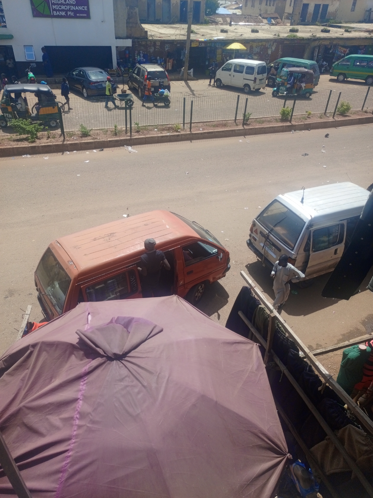 This image shows a street scene in Bukuru, Nigeria, with various vehicles and people present. In the foreground, a large, crumpled purple tarp dominates the view, obscuring part of the scene below. Behind the tarp, an orange van is parked, and a person in a dark shirt is visible through the open driver's side door, appearing to interact with someone inside. To the right of the orange van, a white van is parked with its rear door open, and a person wearing light-colored clothing is standing near it.
Further down the street, several other vehicles are visible. These include a green minivan, a smaller white van, and a tuk-tuk (three-wheeled vehicle) with passengers. A dark sedan is parked on the left side of the street. The background reveals buildings lining the street, some appearing to be shops with awnings and displays. The street itself is paved, and there are various items scattered on the ground, suggesting commercial activity and possibly a market. A metal fence runs along the edge of the sidewalk, separating the street from the buildings. The overall impression is of a busy urban environment with transportation and commerce taking place.