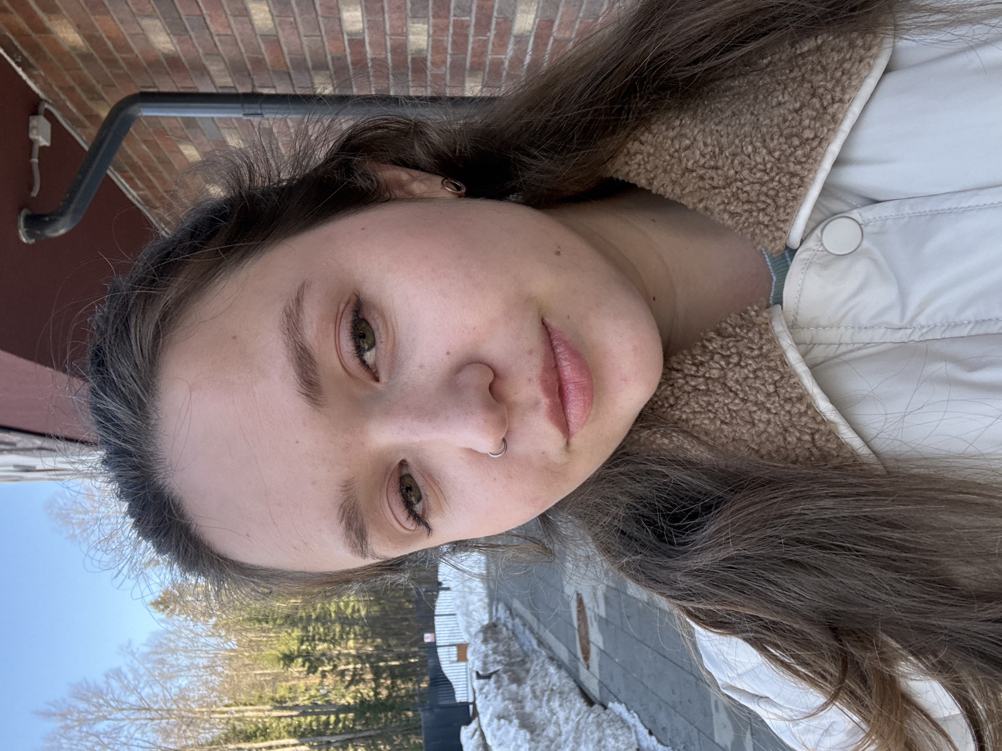 A young woman poses for a close-up selfie. She has fair skin, dark eyebrows, long brown hair, and a nose ring. She wears a light-colored jacket with a sherpa collar. The background shows a brick wall, a pipe, trees, snow, and a paved area. There is no information available to confirm or deny that the picture was taken in the city of Izhevsk, Russia.