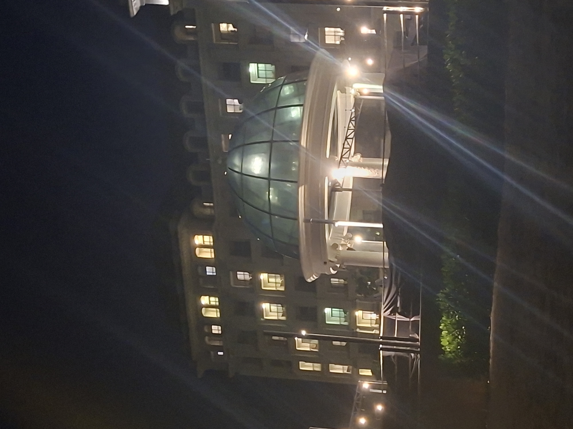 The image captures a nighttime exterior scene of a building resembling a hotel or resort, illuminated by artificial lights. The architecture features multiple stories with numerous windows, many of which are lit, suggesting occupancy. A prominent feature is a glass dome structure in the foreground, possibly a conservatory or an entrance to a facility.
The setting appears to be in a tropical or subtropical location, given the presence of lush green foliage in the lower part of the image. The overall ambiance is one of an established establishment, potentially a tourist destination.
While the image does not provide direct clues to pinpoint a specific location within Phú Quốc, Vietnam, the style of architecture and the tropical vegetation are consistent with the characteristics of resorts and hotels found on the island. The scene depicts a static environment, with no individuals or specific activities clearly visible. The lighting suggests it is after sunset.