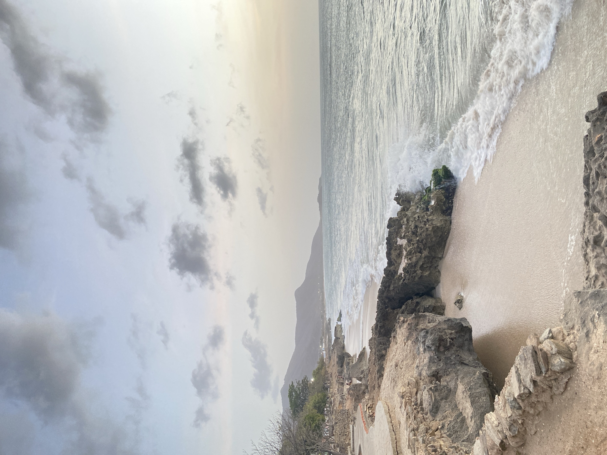The image shows a rocky coastline in Juan Griego, Venezuela, with the ocean waves crashing onto the shore. The sky is overcast, with visible clouds. Mountains are seen in the distance. There are visible manmade structures near the shore. The scene captures a coastal landscape, focusing on the interplay between land and sea, with geological formations and atmospheric conditions present.
