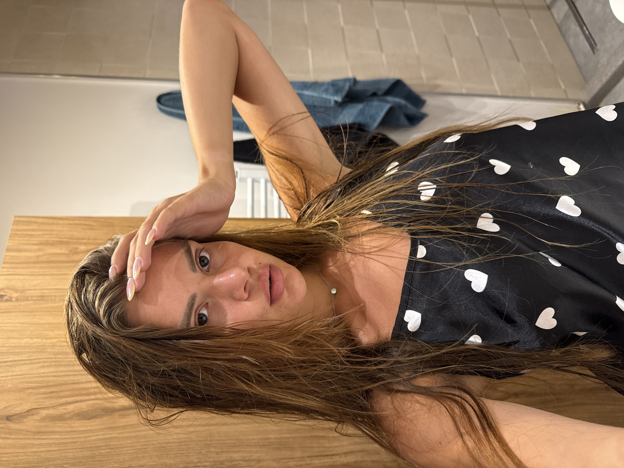 A young woman with wet, long, brown hair is looking at the camera, with her right hand on her forehead. She is wearing a black camisole with white hearts. She has a silver necklace with a small pendant. The background appears to be a bathroom with a wooden cabinet on the left, a tiled wall and shower area on the right, and a towel hanging on a rack. The image was taken in Plovdiv, Bulgaria.