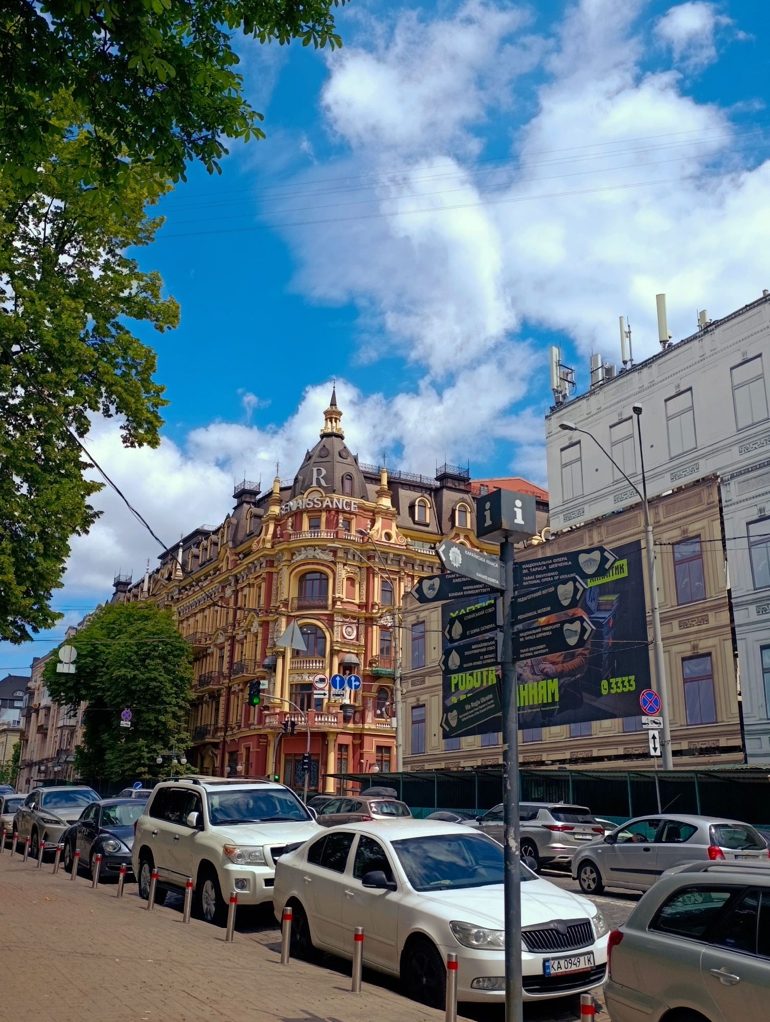 The image shows a street scene in Kyiv, Ukraine, with several cars parked along the side of the road. A large, ornate building with a distinctive domed roof and the name "RENAISSANCE" visible on it dominates the background. The building is painted in shades of yellow and red. To the right of this building, another building is visible, which appears to be more modern and has several antenna-like structures on its roof. In the foreground, a signpost with multiple directional signs points to various locations, including "NATIONAL OPERA OF UKRAINE" and "PEDAGOGICAL MUSEUM." The sky is blue with scattered white clouds. Trees with green foliage are present on the left side of the image. The overall impression is a bustling urban environment with a mix of historical and modern architecture.