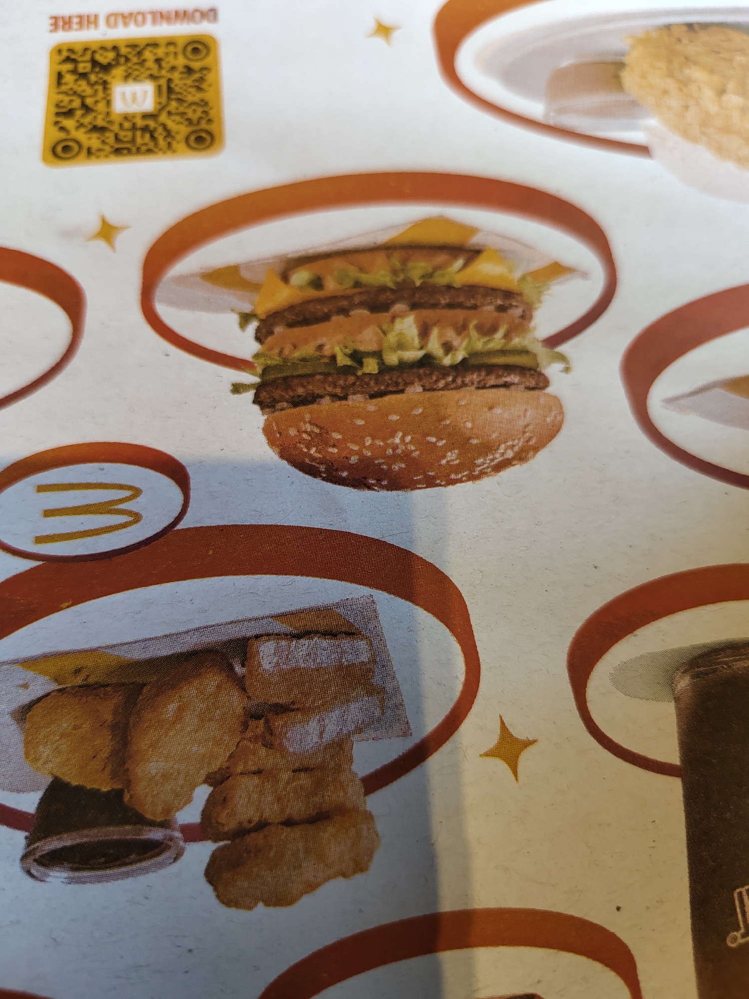 The image shows a menu or promotional material featuring a burger and chicken nuggets, likely from McDonald's, given the visible "M" logo on a white circle with a red border. The burger is a double-patty hamburger with lettuce, cheese, and pickles. The chicken nuggets are golden brown and served with a dipping sauce. A QR code with a white "W" logo is also visible, along with the text "DOWNLOAD HERE". The background is white with orange circular accents and yellow star shapes. The image does not provide any information about a specific location in Davao City, Philippines.