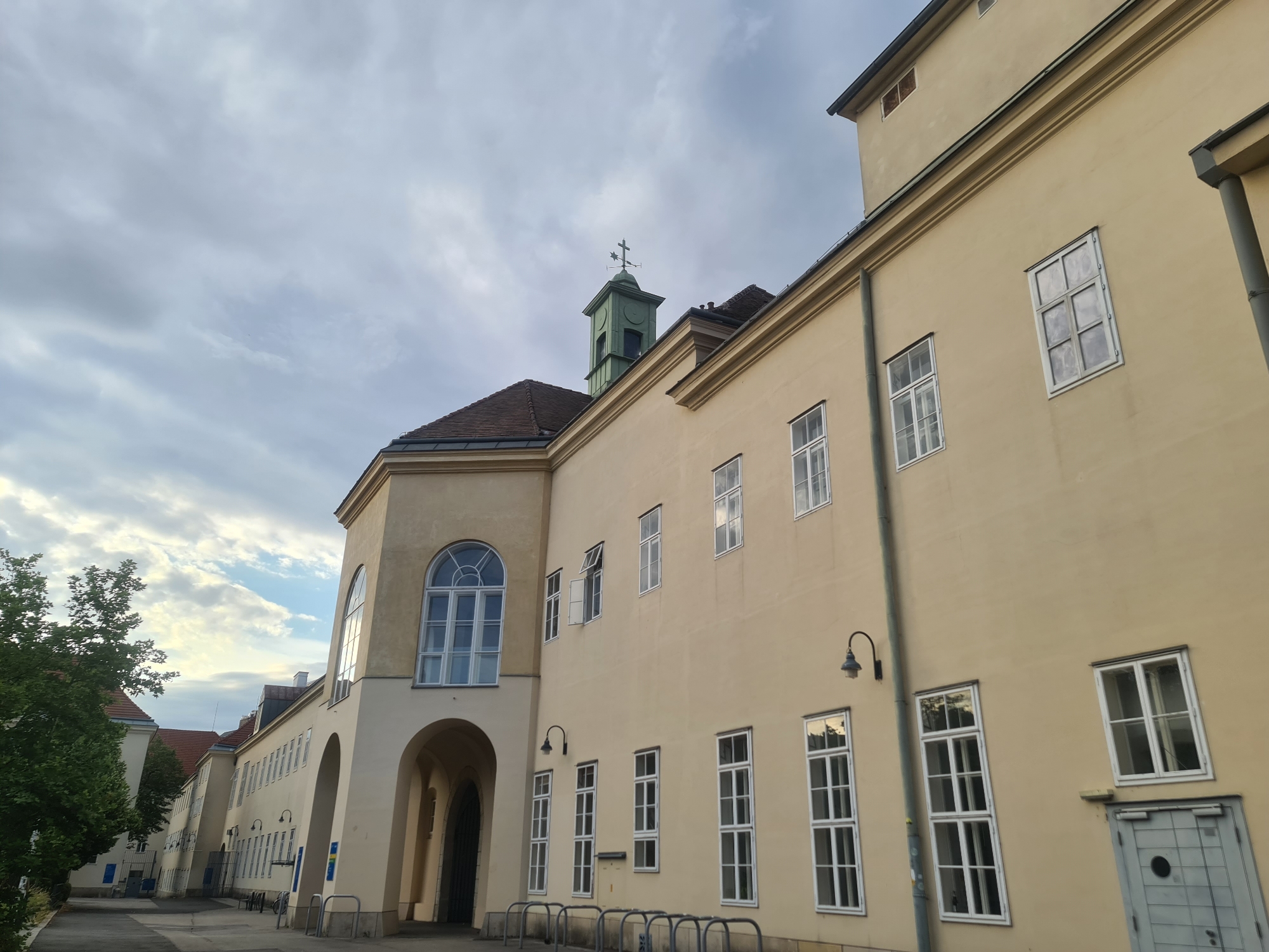 The image shows the exterior of a pale yellow building, possibly an educational institution, with a green bell tower topped by a cross and weather vane, located in Vienna, Austria. The building features numerous windows, some with arched tops, and an arched entryway. A paved pathway runs alongside the building, with trees and other buildings visible in the background. The sky is overcast with scattered clouds. There are no people visible in the scene.