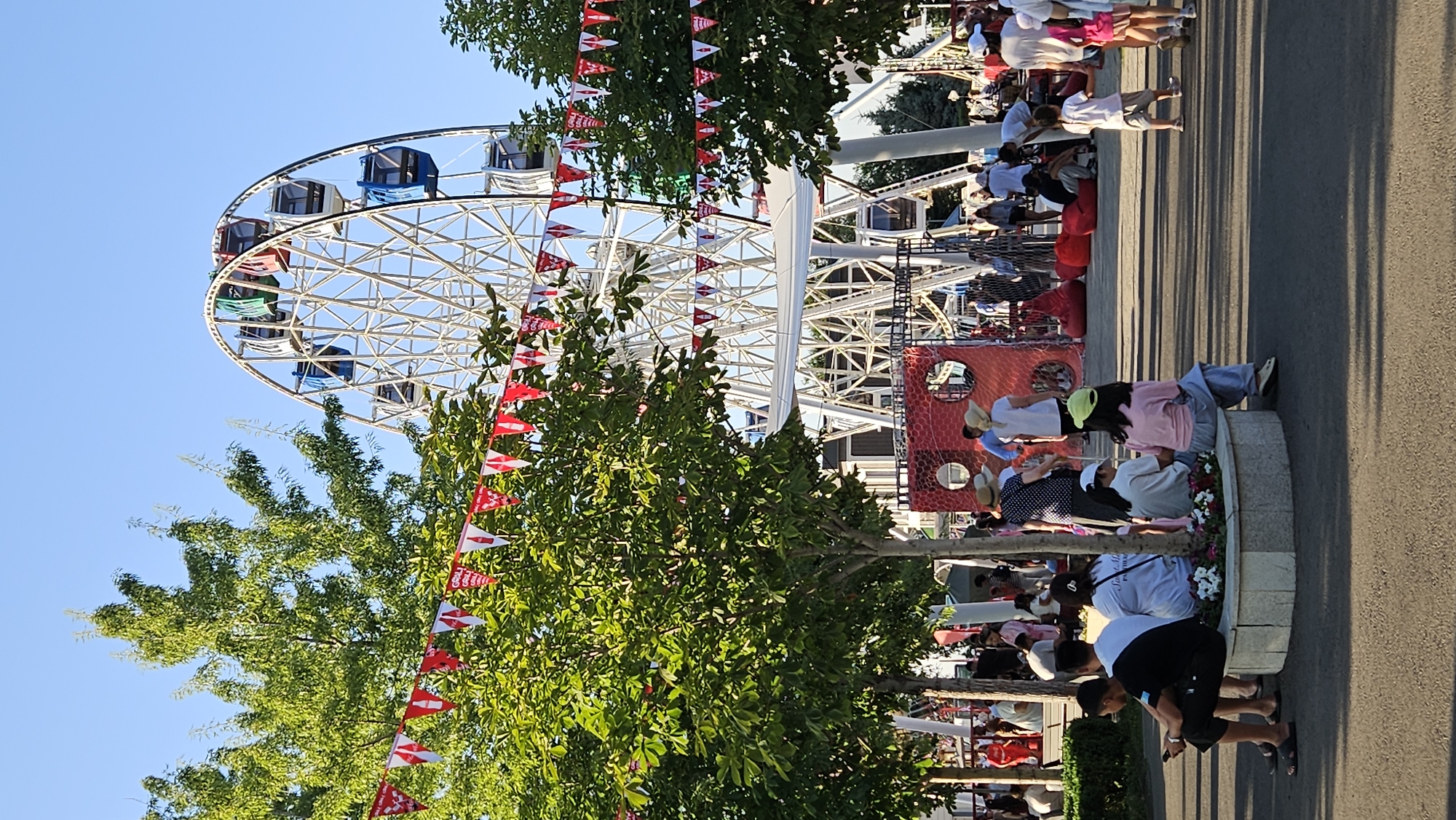 The image depicts a sunny day at an amusement park, possibly in Almaty, Kazakhstan, as suggested by the presence of a Ferris wheel and numerous families enjoying outdoor leisure activities. In the foreground, several people are seated on a circular, stone-edged planter with flowers, facing away from the camera. Behind them, a red play structure with circular openings is visible, along with red beanbag chairs scattered on the ground. A large Ferris wheel dominates the background, with some of its gondolas in motion. Red and white triangular flags are strung between trees and poles, adding a festive atmosphere. People of various ages are scattered throughout the park, some walking, some sitting, and some standing. The environment appears to be a paved area with scattered trees, casting long shadows on the ground. The sky is clear and blue, indicating good weather.