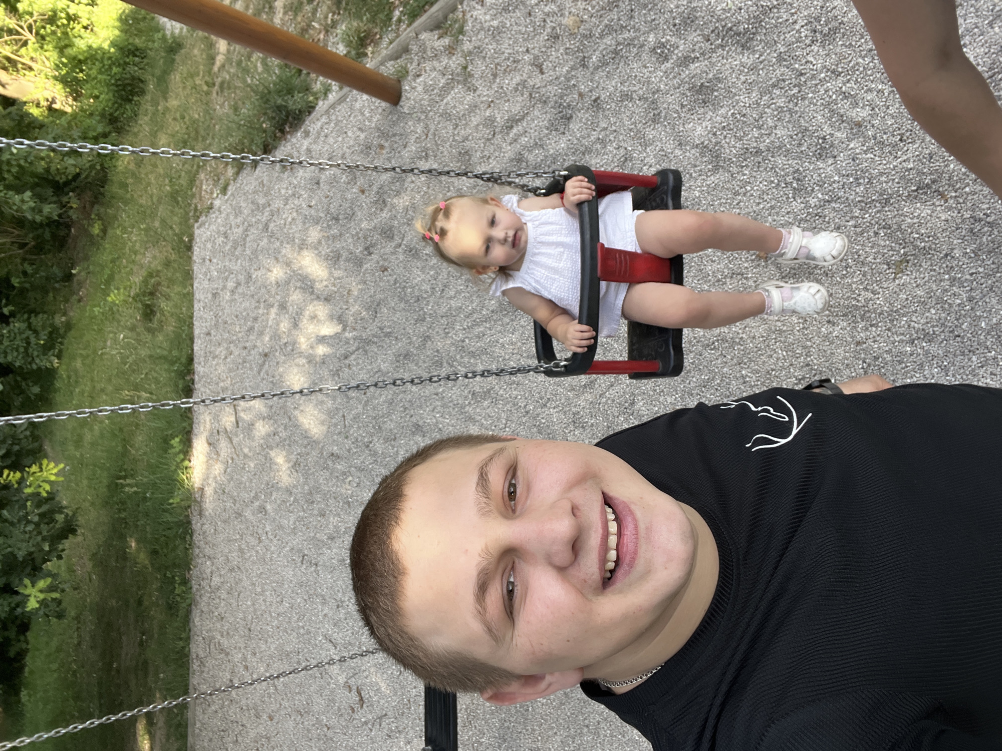 A young man and a toddler are on a playground in Pilsen, Czechia. The man is in the foreground, smiling directly at the camera, wearing a black, ribbed t-shirt with a white line drawing on the left chest. A silver chain is visible around his neck. Behind him, a toddler is seated on a red and black swing, wearing a white textured dress and white sandals with pink accents. The swing set is suspended by chains. The playground surface is covered in light-colored gravel. In the background, trees and green grass are visible. A wooden support beam for the swing set is on the right side of the frame. The lighting suggests it is daytime.