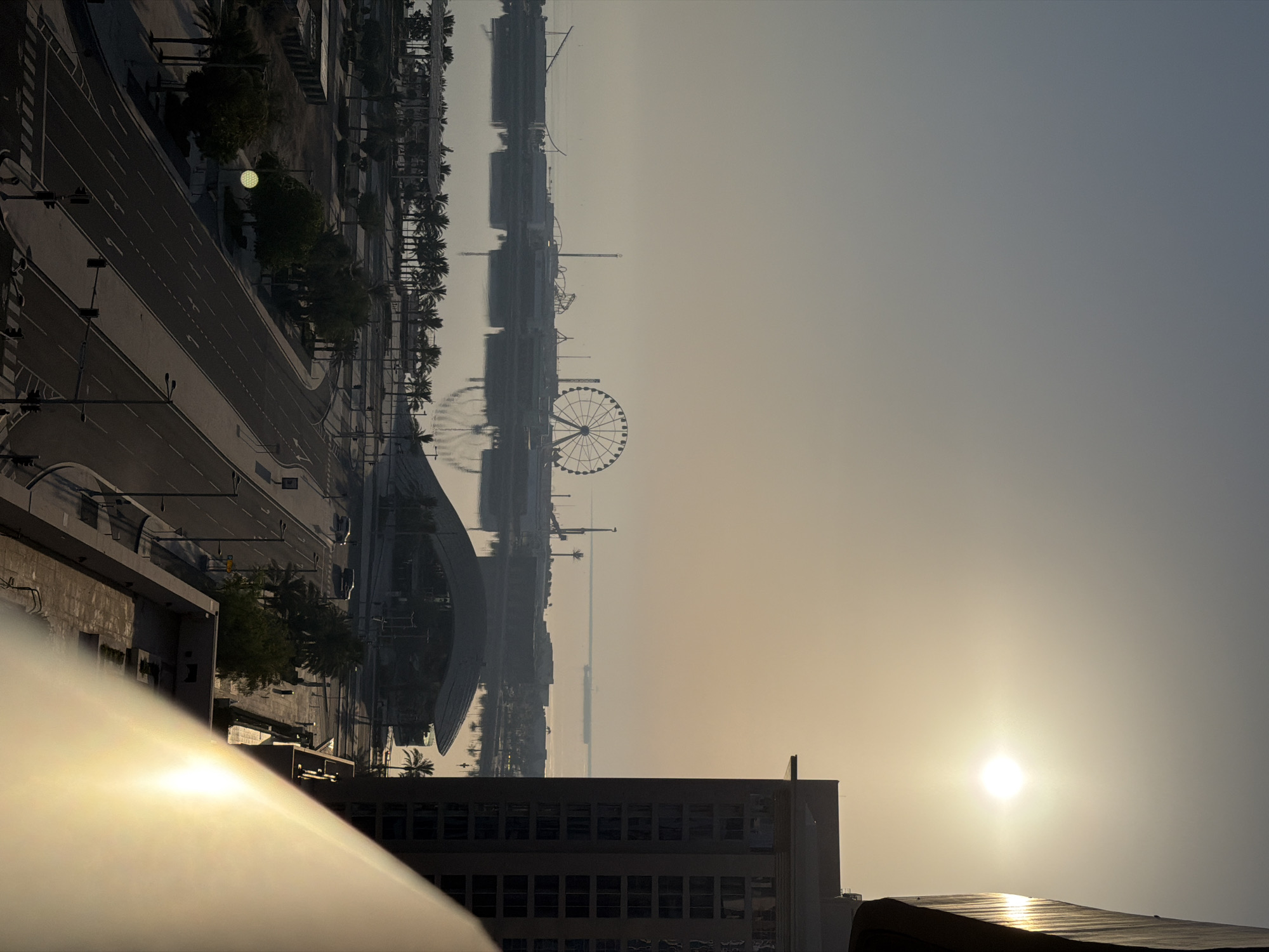 The image shows an aerial view of a city waterfront during a bright sunrise. A large Ferris wheel is prominent on the left side of the image, silhouetted against the hazy sky. In the foreground, a modern building with many windows is visible on the right, with a balcony railing in the lower right corner reflecting the sunlight. A road with traffic signals and palm trees lines the waterfront. A body of water reflects the structures and palm trees. The overall scene suggests an urban environment with recreational attractions.