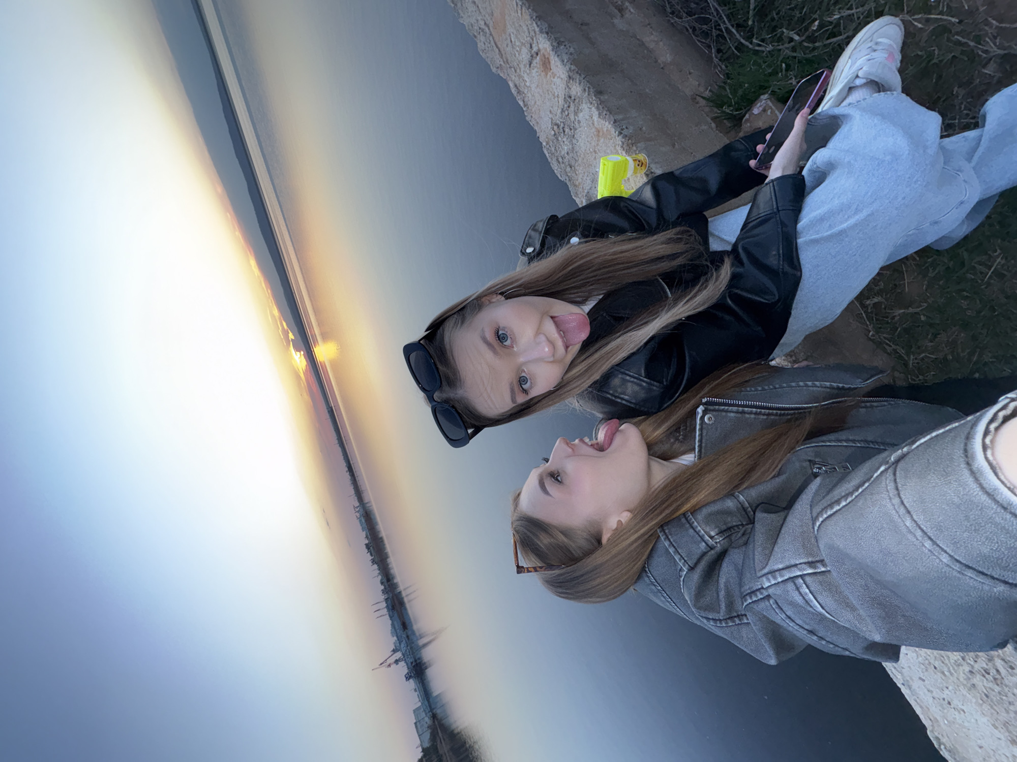 Two young women are posing for a selfie on an elevated structure, possibly a bridge or embankment, overlooking a large body of water at sunset. The woman on the top is wearing a black leather jacket, light blue jeans, and white sneakers; she is holding a cell phone. The woman on the bottom is wearing a gray denim jacket. Both women are sticking their tongues out playfully. The background shows a body of water reflecting the colors of the setting sun. In the distance, there are some indistinct structures which indicate that it is the city of Chaikovsky, Russia.