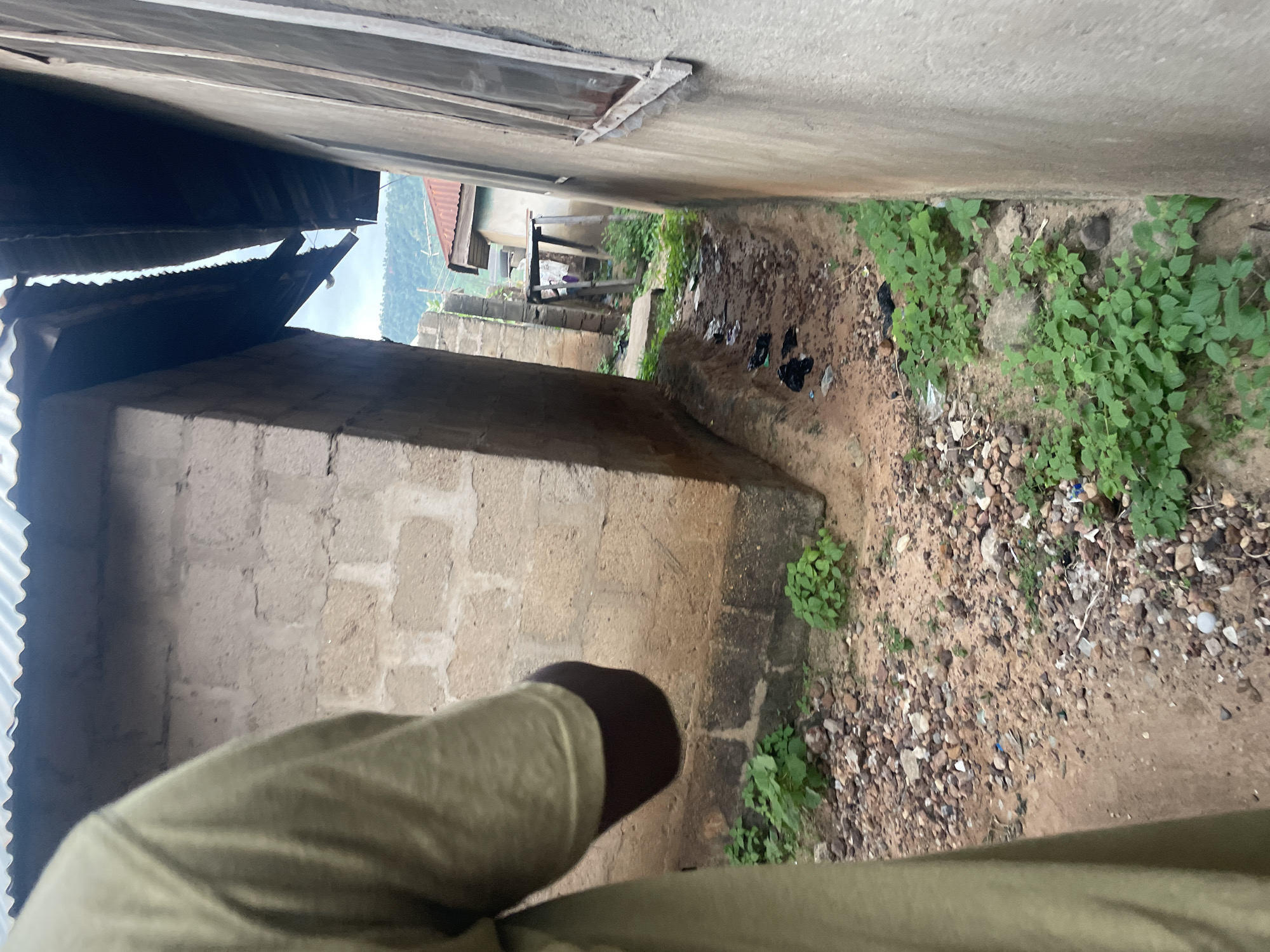 The image depicts a narrow pathway between two buildings, likely located in Lokoja, Nigeria. A person, wearing a light green long-sleeved shirt, is partially visible on the left side of the frame, their arm and shoulder in focus. The pathway is unpaved and covered with gravel, small rocks, and patches of green vegetation. Litter, including black plastic bags, is scattered along the ground.
The building on the left is constructed with concrete blocks and has a corrugated metal roof. The building on the right has a rough concrete exterior and a window with visible wooden frames. In the background, other structures with corrugated metal roofs are visible, suggesting a residential or densely populated area. The overall environment appears to be an urban or semi-urban setting, possibly a back alley or a side street. The lighting suggests it is daytime, with soft natural light illuminating the scene.