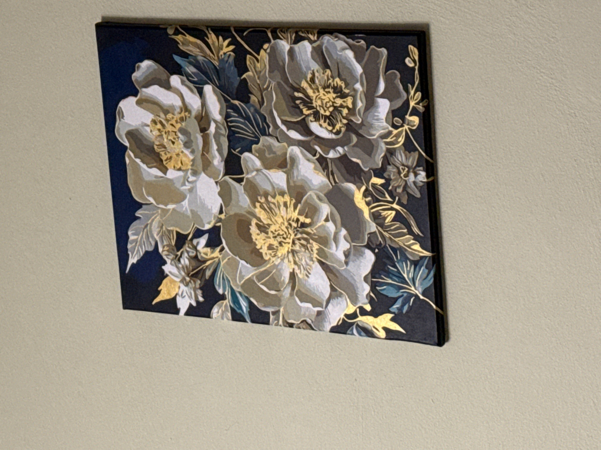 A painting of three large, light-colored flowers with yellow centers and gold accents is displayed on a light beige wall. The painting has a dark navy background and features green and gold leaves and vines. The painting is hung at a slight angle, tilted to the left. The wall texture is visible and appears to be a rough, matte finish. The lighting in the scene is even, with no harsh shadows cast by the painting or any other objects. The overall impression is one of a decorative art piece in a simple domestic setting.