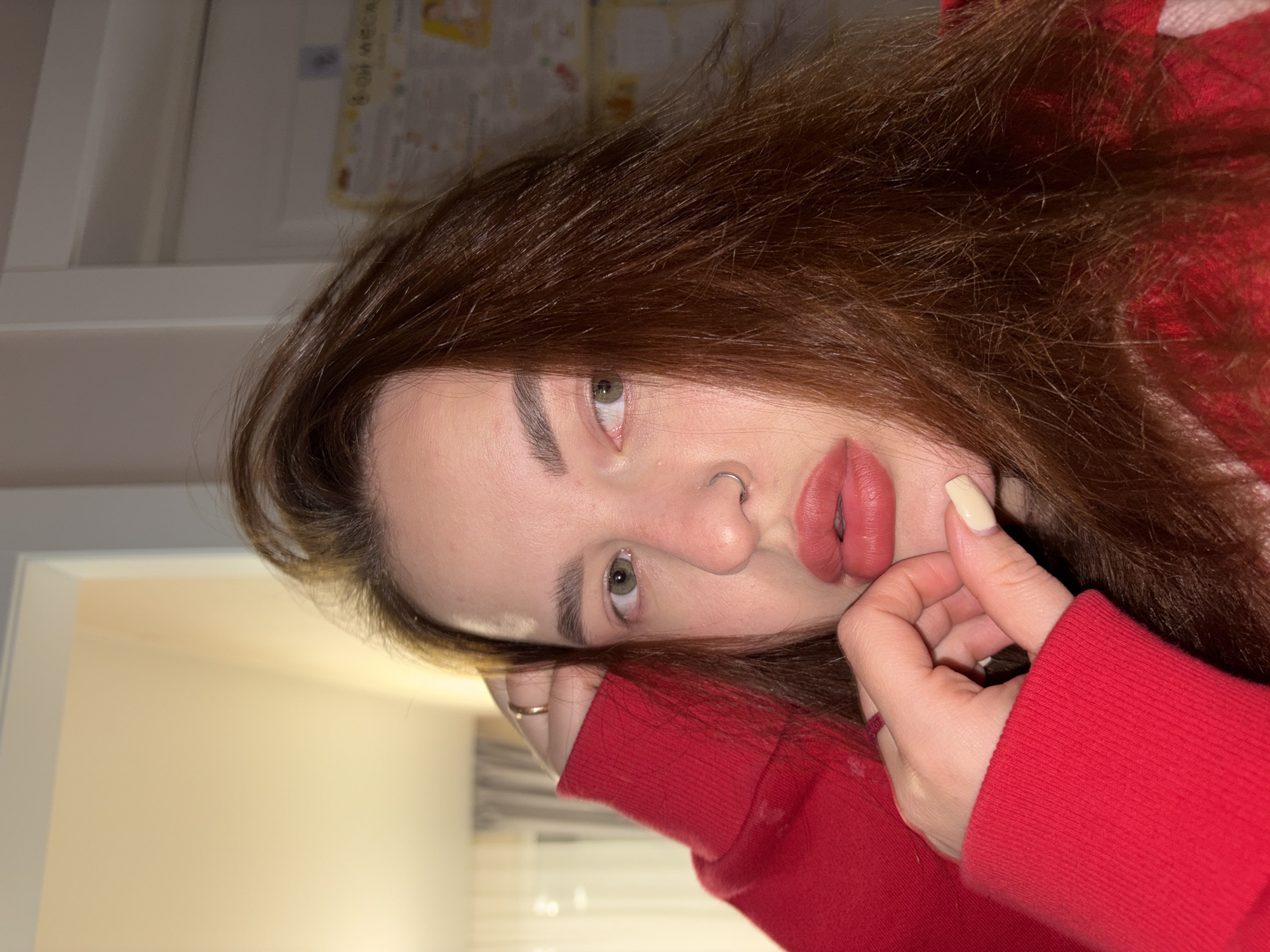 A close-up, rotated portrait features a young woman with long, reddish-brown hair. She is wearing a red long-sleeved top or sweatshirt. Her left hand, adorned with a gold-colored ring on the ring finger, rests near her chin and mouth, with one visible nail painted a light cream color. She has a silver septum piercing. Her eyes are a light green/hazel, and her lips are full and subtly parted, colored with a rose-toned lipstick. She maintains direct eye contact with the viewer.
The setting is indoors, appearing to be a residential room, indicated by the plain light-colored walls and ceiling/upper wall section. In the blurred background, specifically on the upper right, a light-colored chart or poster with some indistinct Cyrillic text is visible, hinting at the location context of Krasnodar, Russia. The lighting suggests an indoor environment, likely illuminated by artificial light. The overall mood is calm and direct.