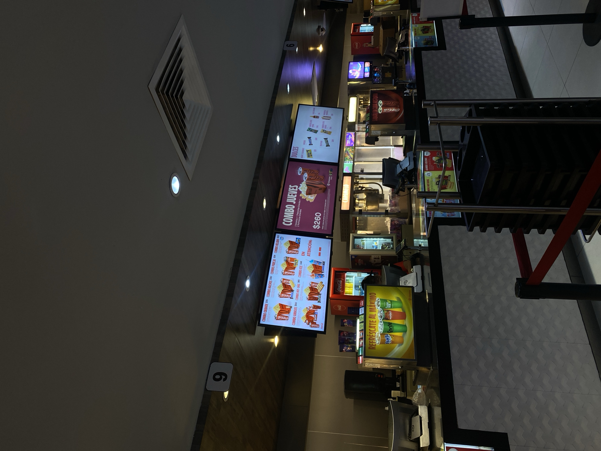 The image displays a brightly lit concession stand, likely situated within a cinema or entertainment complex, captured from an elevated, rotated perspective. The scene focuses on the serving area with a variety of food and beverage options.
In the upper left, a large square air vent is visible on a light grey ceiling, accompanied by a single recessed light. A wall transitions from light grey to a wooden paneling effect, featuring several small recessed lights and a white sign with the number "6".
The central focus is a series of three vertical digital menu boards. The leftmost board, labeled "DULCES" (Sweets/Candies), lists various snack items. The middle board advertises a "COMBO JUEVES" (Thursday Combo) for "$260," which includes "2 Refrescos Grande" (2 large sodas) and "2 Boletos + 1 Palomitas Grandes" (2 tickets + 1 large popcorn). Other options like "COMBO GRANDE $275," "COMBO NACHOS," "COMBO PAREJA," and "COMBO HELADO" are also displayed. The rightmost menu board shows "COMBO AMIGOS $563," "COMBO HOT-DOGS," and "COMBO ICEE," with images of the respective items and "EN ATENCIÓN:" (At the counter/In attendance).
To the right of the menu boards, a horizontal illuminated sign promotes "REFRESCATE AL MÁXIMO" (Refresh yourself to the maximum), featuring images of Fanta, Sprite, Mundet, and Coca-Cola. Behind a counter, a staff member is partially visible, operating the station with Coca-Cola branded beverage dispensers and coolers in the background. Queuing barriers, colored black with red straps, are positioned in the foreground on the right, indicating a customer line. The floor is covered with patterned grey tiles. Another white sign with the number "6" is visible in the lower right, potentially marking a station or queue number. The overall setting suggests a busy, modern snack bar environment.