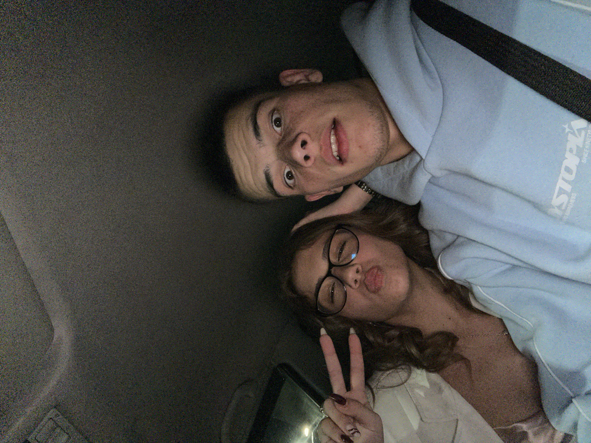 Two individuals are in what appears to be the interior of a car. A young man is wearing a light blue hoodie, and a seatbelt is visible across his chest. A young woman is wearing glasses and a light-colored top, flashing a peace sign with her right hand. The interior appears dark. The location in Karavelovo, Bulgaria is not determinable.