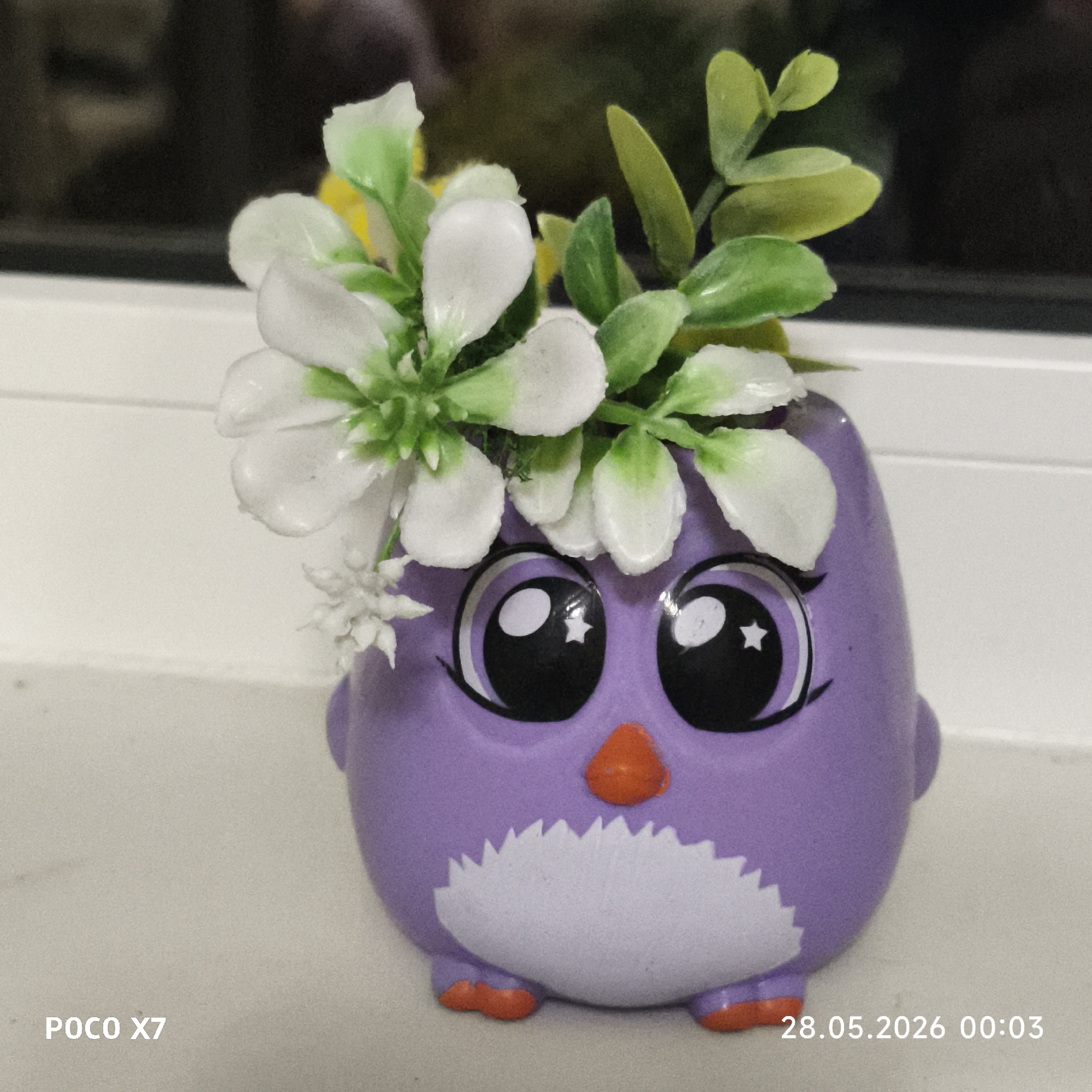 A purple owl figurine with large, cartoonish eyes and a small orange beak is situated on a white surface. White flowers with green accents are arranged on top of the figurine's head, resembling a headdress. Green leaves are also mixed in with the flowers. The figurine has a white, scalloped pattern on its chest and small orange feet. The background is out of focus, suggesting a room with a window. The text "POCO X7" and "28.05.2026 00:03" are present in the lower corners of the image.