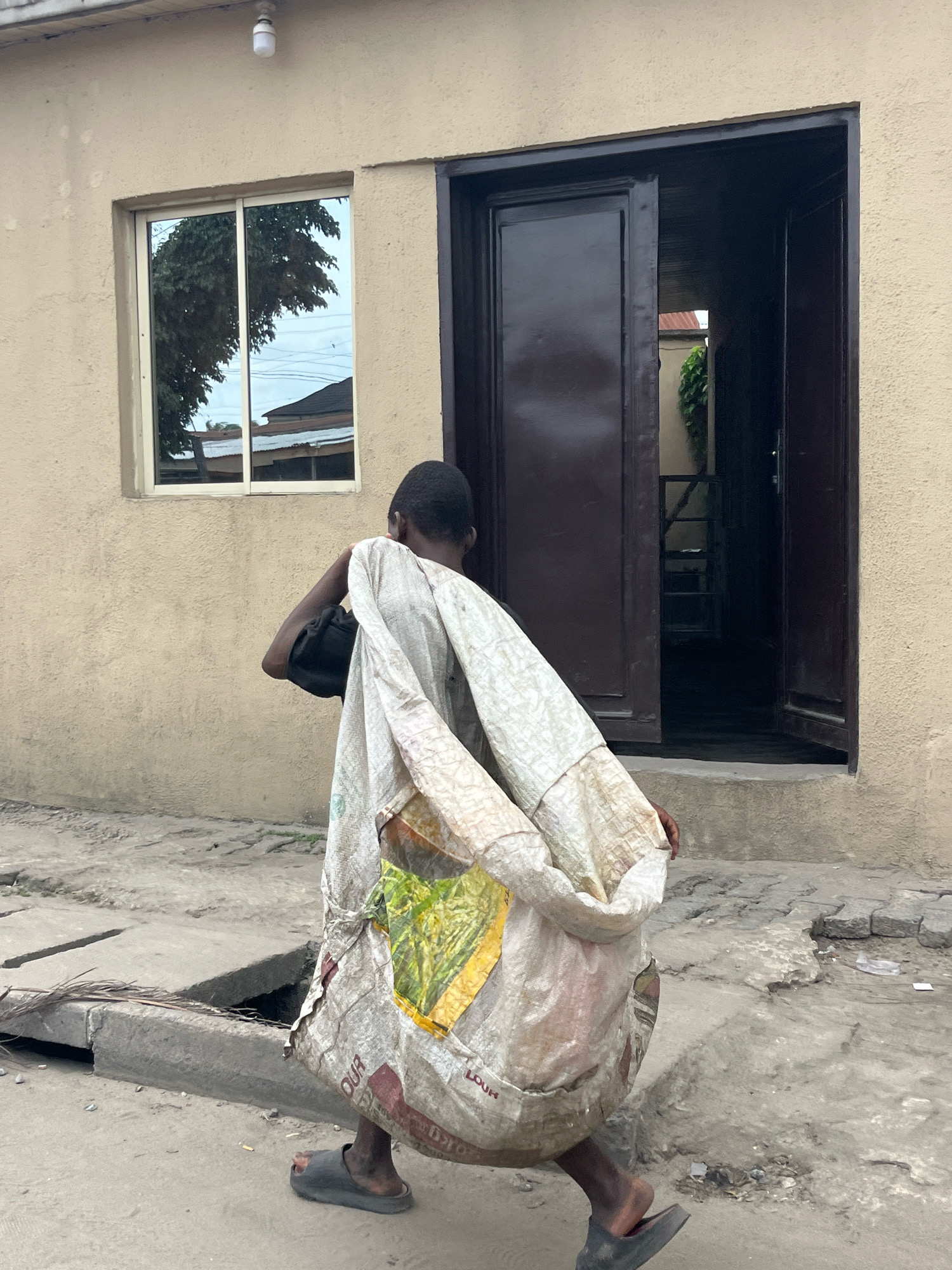 A young person, from the back, walks away from the camera on a paved street in Ado, Nigeria. They are carrying a large, white, roughly woven sack over their right shoulder, with a black bag tucked underneath the sack. The sack appears to be made of repurposed material and has some red and yellow printed text and graphics visible. The person is wearing dark-colored, open-toed sandals.
They are walking past a beige building with a dark brown, double-door entryway that is partially open, revealing a dark interior space with shelves. To the left of the entryway, there is a horizontal, silver-framed window reflecting trees and buildings. The ground is concrete with some visible cracks and debris, and a grate is present on the left side of the street. The lighting suggests it is daytime.