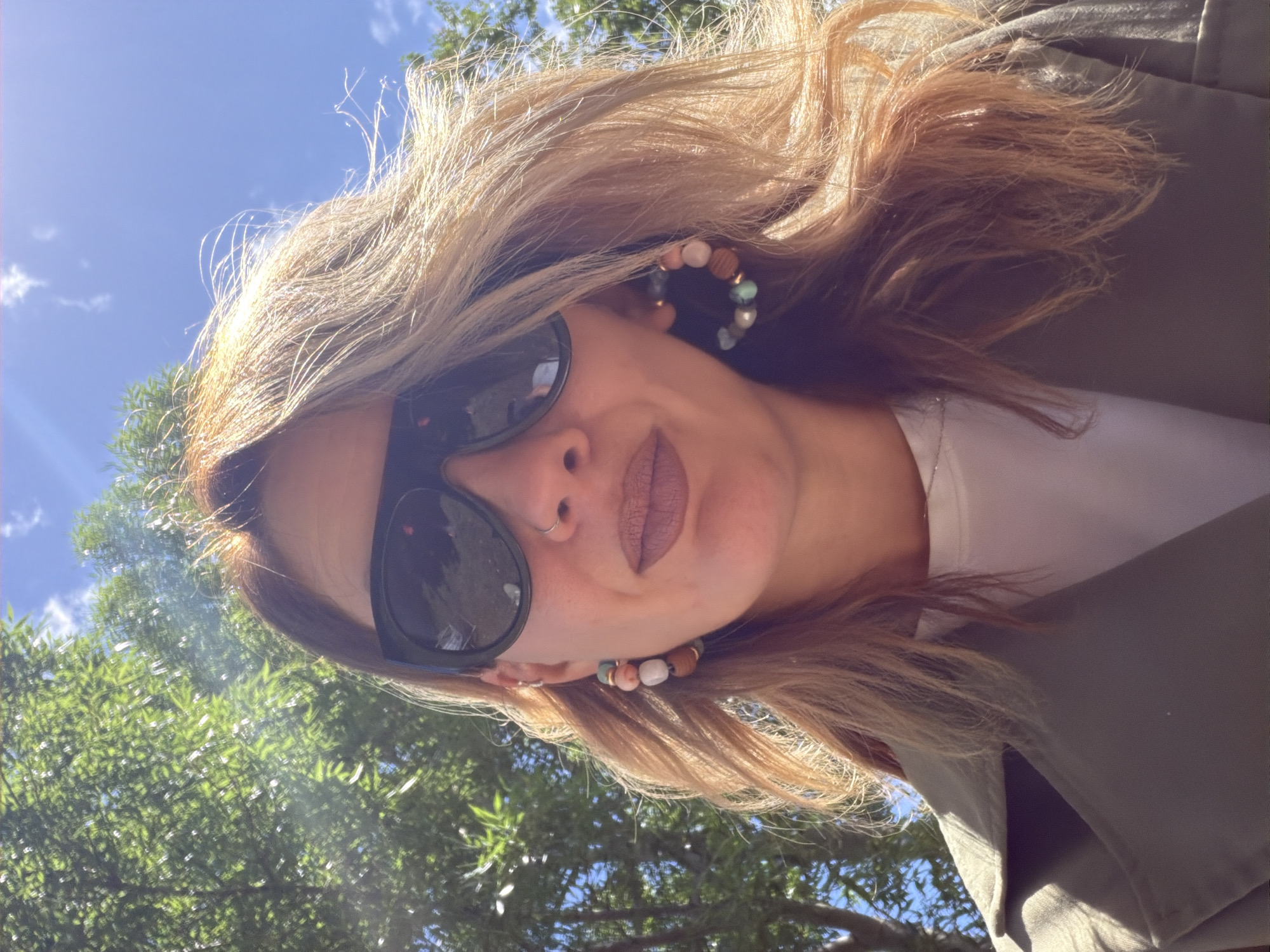 A woman with long, blonde hair, sunglasses, and hoop earrings takes a selfie outdoors. She is wearing a white t-shirt and a green jacket. In the background, there are green trees and a blue sky with some clouds. The image does not contain information that places it in the city of Epsom and Ewell, United Kingdom.
