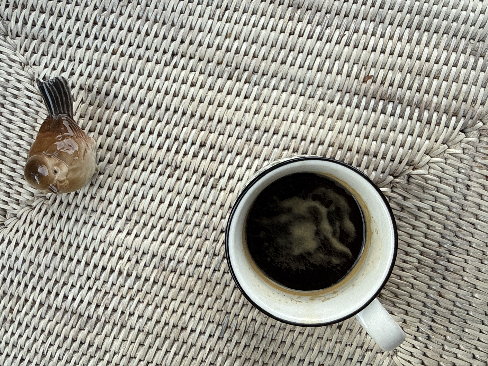 A ceramic bird figurine is placed on a woven textured surface, to the right of a white mug filled with dark coffee. The mug has a black rim and a black stripe around its base. The coffee has a frothy layer on top. The woven surface is light brown and appears to be made of natural fibers, possibly rattan or bamboo, and is laid out on a flat surface such as a table or chair. The scene is lit from above, casting subtle shadows. The bird figurine is light brown with darker brown and black markings on its tail feathers, and it has small dark eyes. The surface and objects suggest an outdoor or patio setting. The image provides no information about the city of Bad Sachsa, Germany.