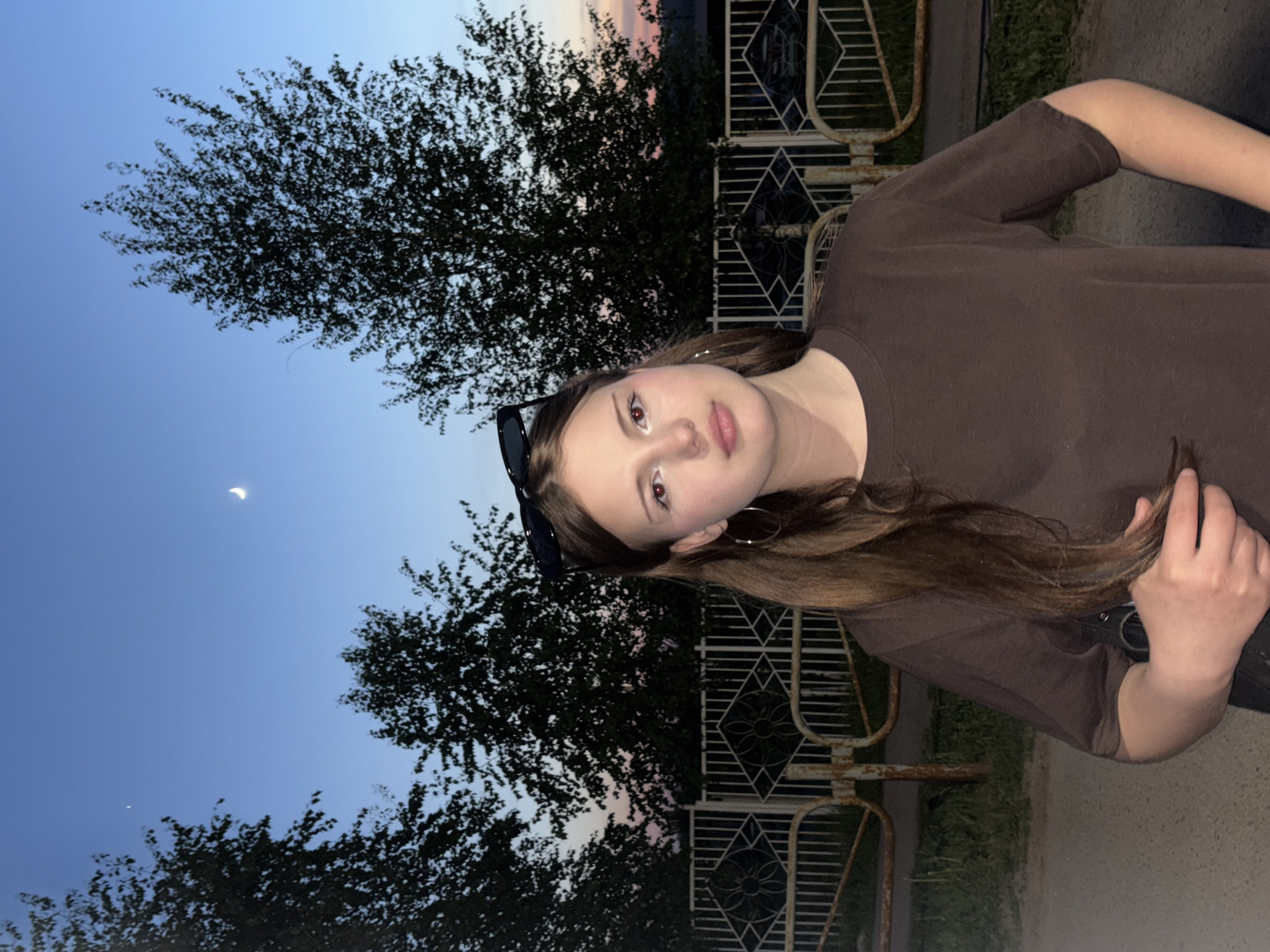 A medium shot of a young person with long brown hair and fair skin, wearing a brown t-shirt and hoop earrings. Sunglasses rest on top of their head. The subject is standing outdoors near a metal fence with a decorative diamond pattern. Behind the fence, trees fill the background beneath a twilight sky with a thin crescent moon visible. The image location is likely in Фоки, Russia.
