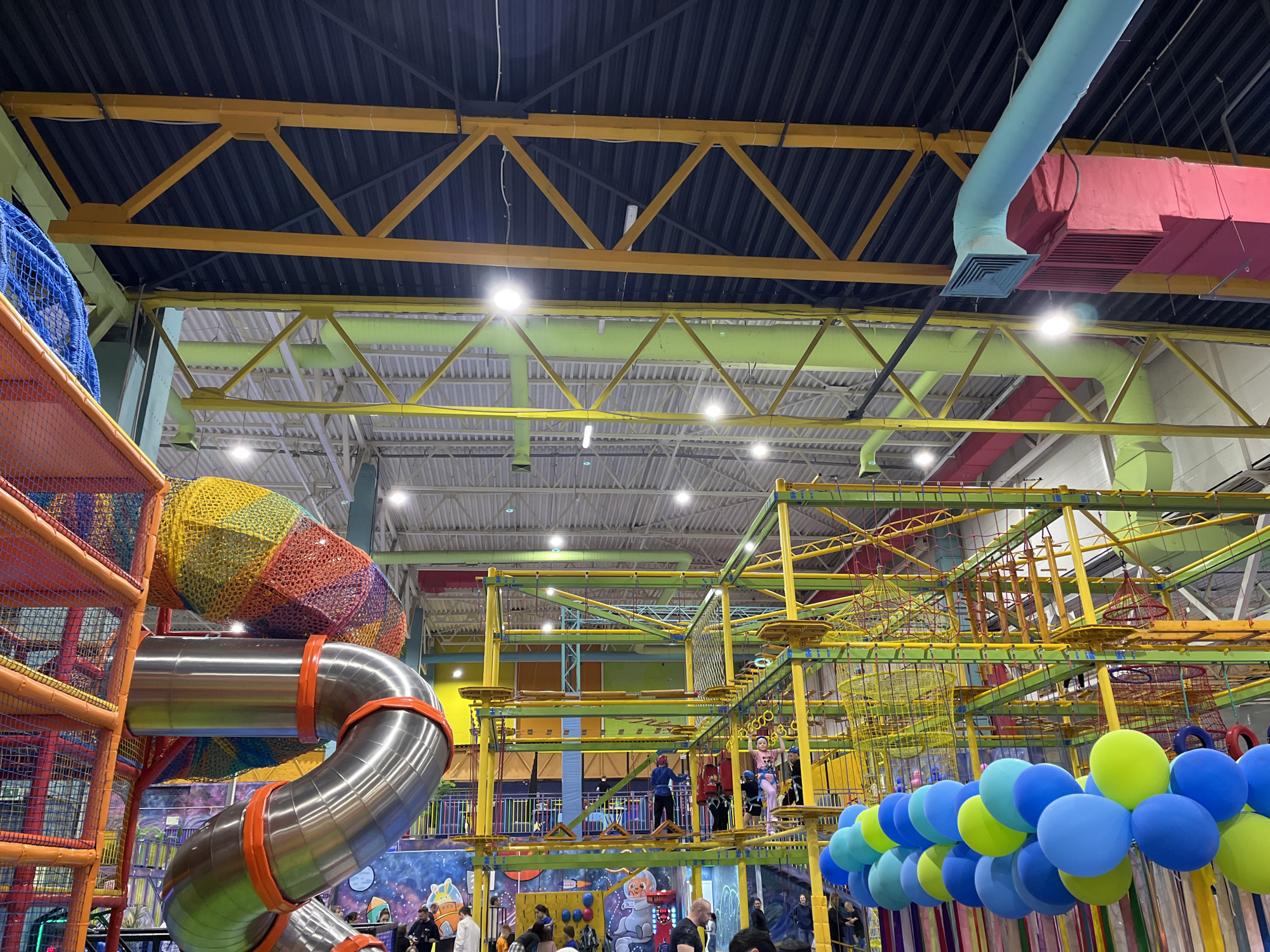 The image shows an indoor playground in Krasnoyarsk, Russia. The playground features a large metal slide, climbing structures with netting, and an elevated obstacle course. Blue and green balloons decorate the area. People, including adults and children, are visible playing on the structures and observing from below. The ceiling has a complex arrangement of yellow and green beams and ventilation systems. The walls are decorated with murals depicting space themes.