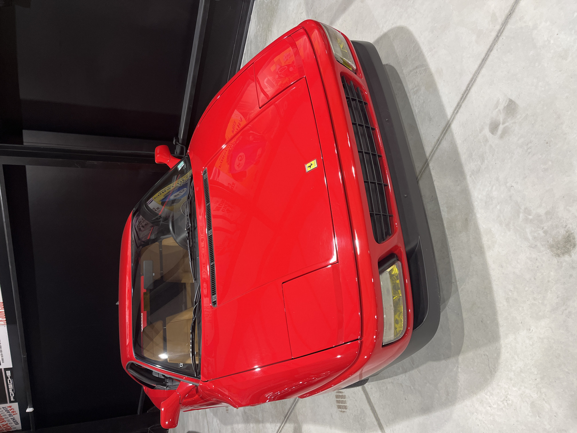 A red Ferrari Testarossa is parked indoors on a light gray floor. The car is angled towards the upper right corner of the image, revealing its front end and passenger side. The Ferrari logo is visible on the hood. The interior is tan. To the left of the car, a black vertical structure is present, with a poster containing the word "Porsche" visible on the left side of the frame. The car is likely indoors, perhaps in a showroom or museum. There is no indication of the location being Kapatovo, Bulgaria.