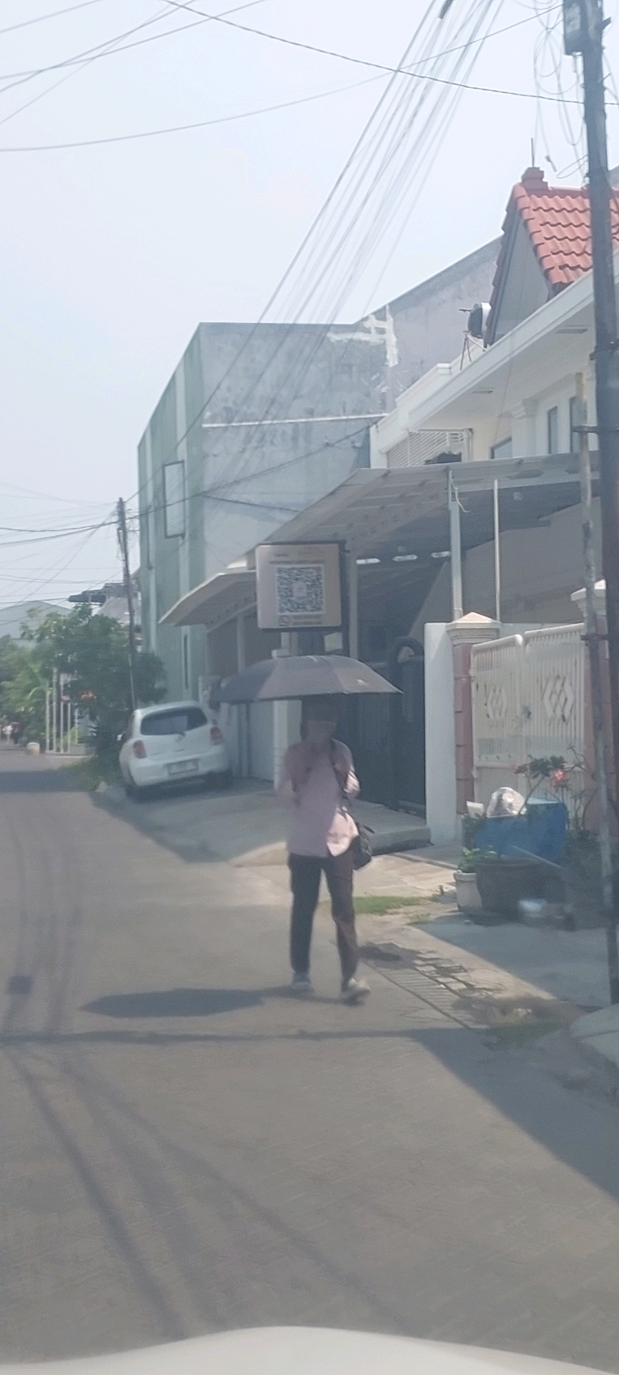 In Surabaya, Indonesia, a person walks down a paved street carrying a black umbrella. The individual is wearing a light pink collared shirt and dark pants. They are also carrying a dark bag slung across their body. To the left of the person, a white car is parked on the side of the road, near a white fence. In the background, a modern building with a red-tiled roof is visible, along with power lines crisscrossing the sky. A sign with a QR code is mounted on a pole in front of the building. The scene appears to be a residential or commercial area on a clear day.