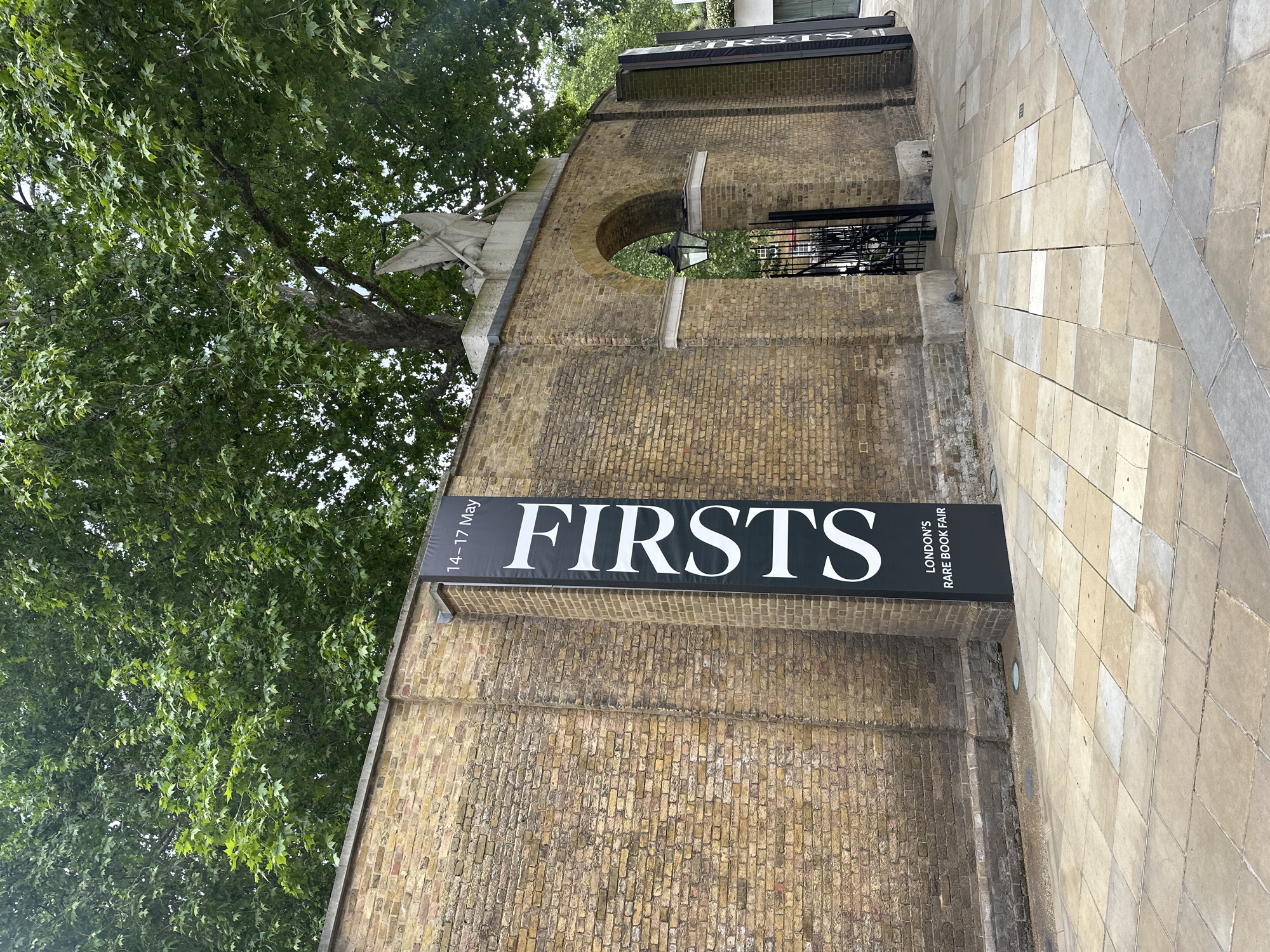 A sideways shot depicts a portion of a brick building featuring a sign that reads "FIRSTS" in bold, white letters, with smaller text below indicating "LONDON'S RARE BOOK FAIR" and dates "14-17 May." An arched entrance with a black metal gate is visible, and foliage from a tree overhangs the structure. The building is located in the city of London, United Kingdom, as indicated on the sign. The foreground includes a section of paved walkway.