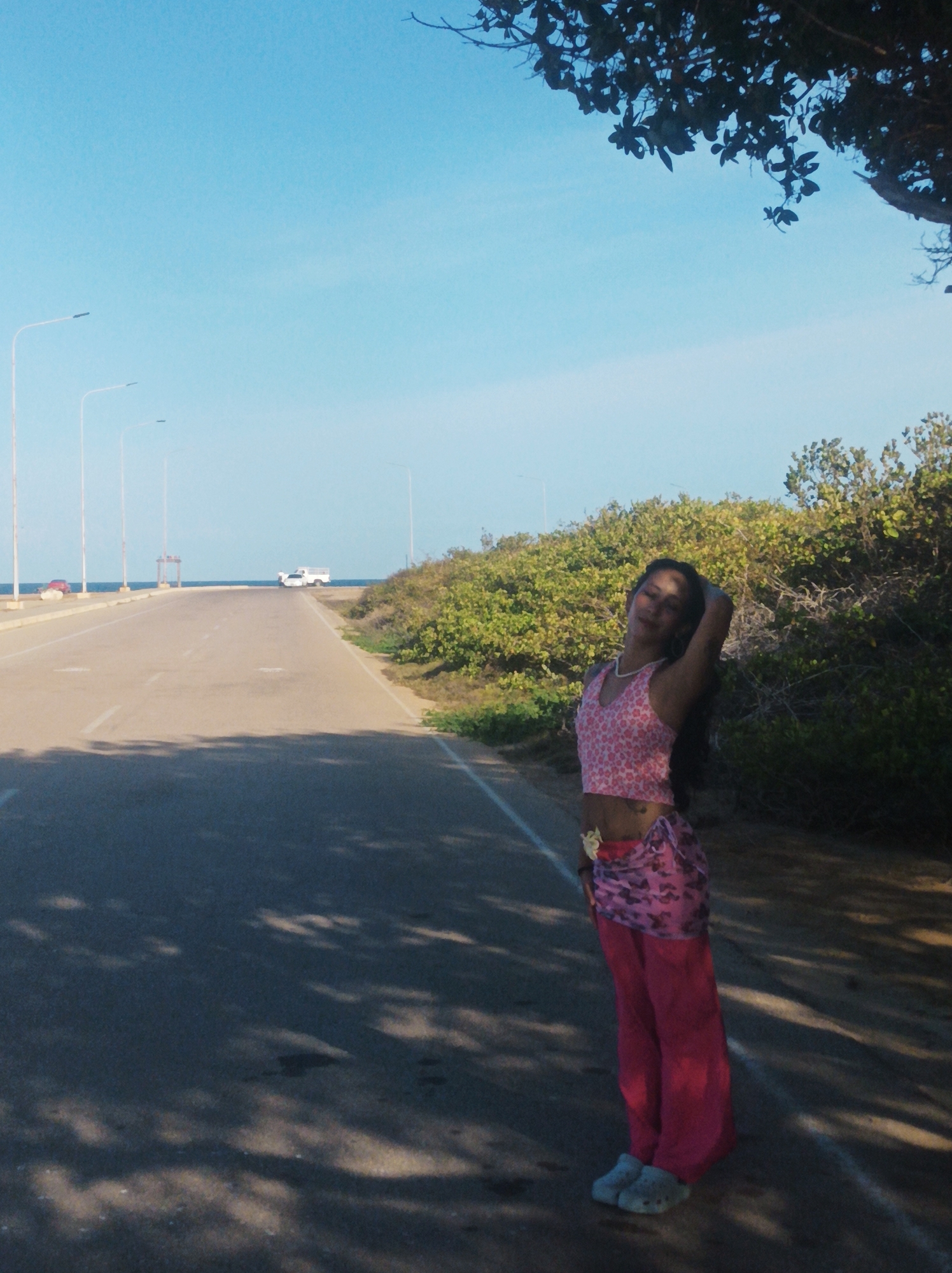 A long straight road with light poles on the left and greenery on the right stretches towards the horizon where the sea is visible in the distance. A light-colored vehicle is parked on the road ahead. A woman stands on the right side of the road, posing with her left hand behind her head. She is wearing a pink crop top, a pink sarong-like garment, and pink pants with light-colored shoes. Shadows of foliage cover the lower part of the road. The sky is clear and blue. The location is likely a coastal road near Porlamar, Venezuela.