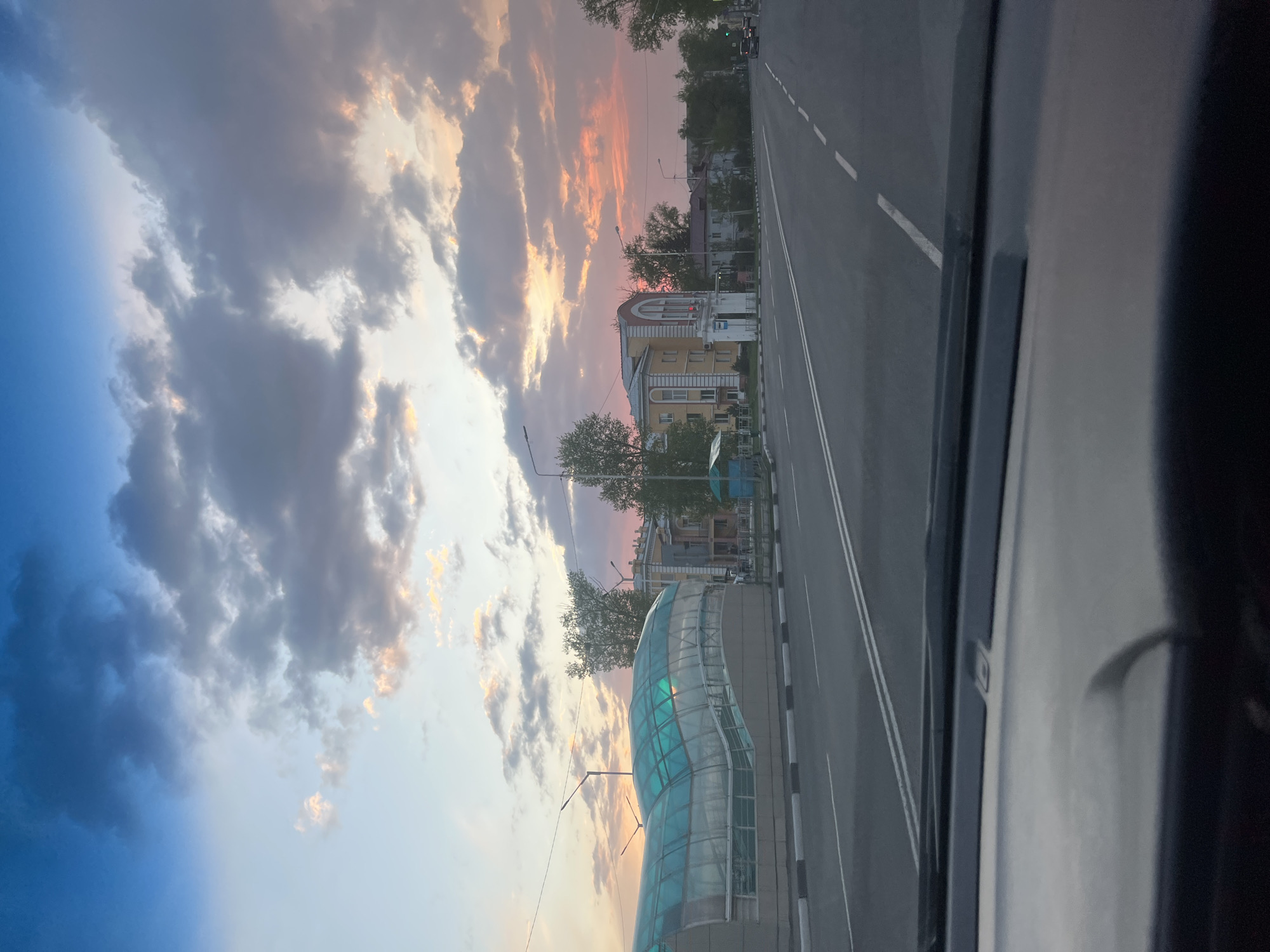 The image depicts an urban scene at sunset, captured from within a vehicle. The view is primarily of a multi-lane road with white dividing lines, flanked by a sidewalk with alternating black and white blocks. Along the road are various buildings, including a two-story yellow building with arched windows and a blue glass-enclosed structure, possibly an overpass or bus stop. Trees are interspersed along the roadside. The sky displays a gradient of colors from orange near the horizon to blue higher up, with scattered clouds. The overall perspective suggests the photo was taken through a windshield from a moving vehicle.