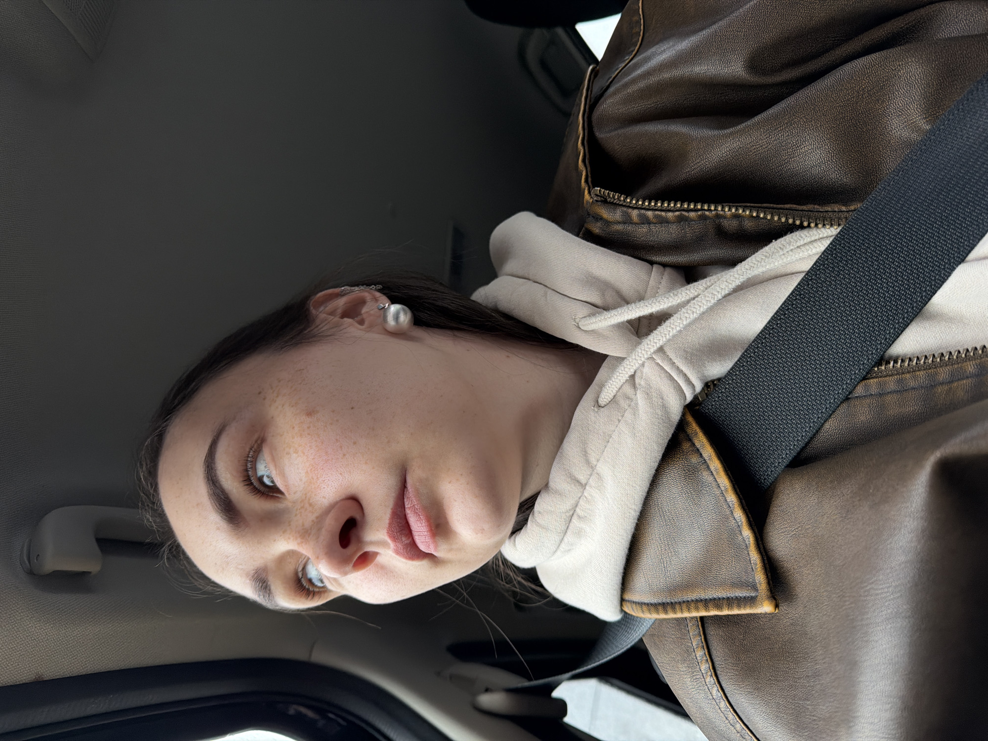A woman is seated in a car wearing a brown leather jacket over a light beige hooded sweatshirt. She has a black seatbelt fastened across her torso. She has light skin with freckles, dark hair, and is wearing a silver earring with a round, pearl-like stud. The interior of the car is visible, including the headliner, a grab handle, and part of the dashboard. The location is not discernible, and there is no indication the photo was taken in Chaikovsky, Russia.