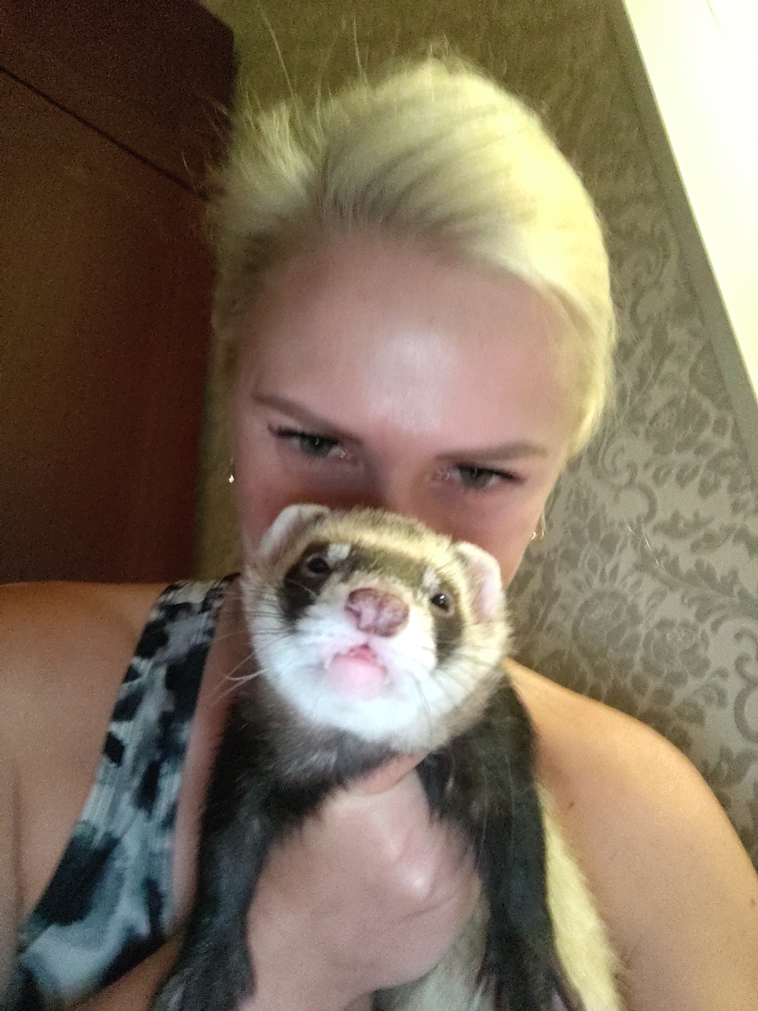 A woman with blonde hair is holding a ferret in front of her face. The ferret's nose and mouth are visible, and it appears to be looking at the camera. The woman is wearing a patterned tank top. The background is out of focus, but appears to be a room with patterned wallpaper. There is no identifiable location in the city of Kyiv, Ukraine visible in this image.