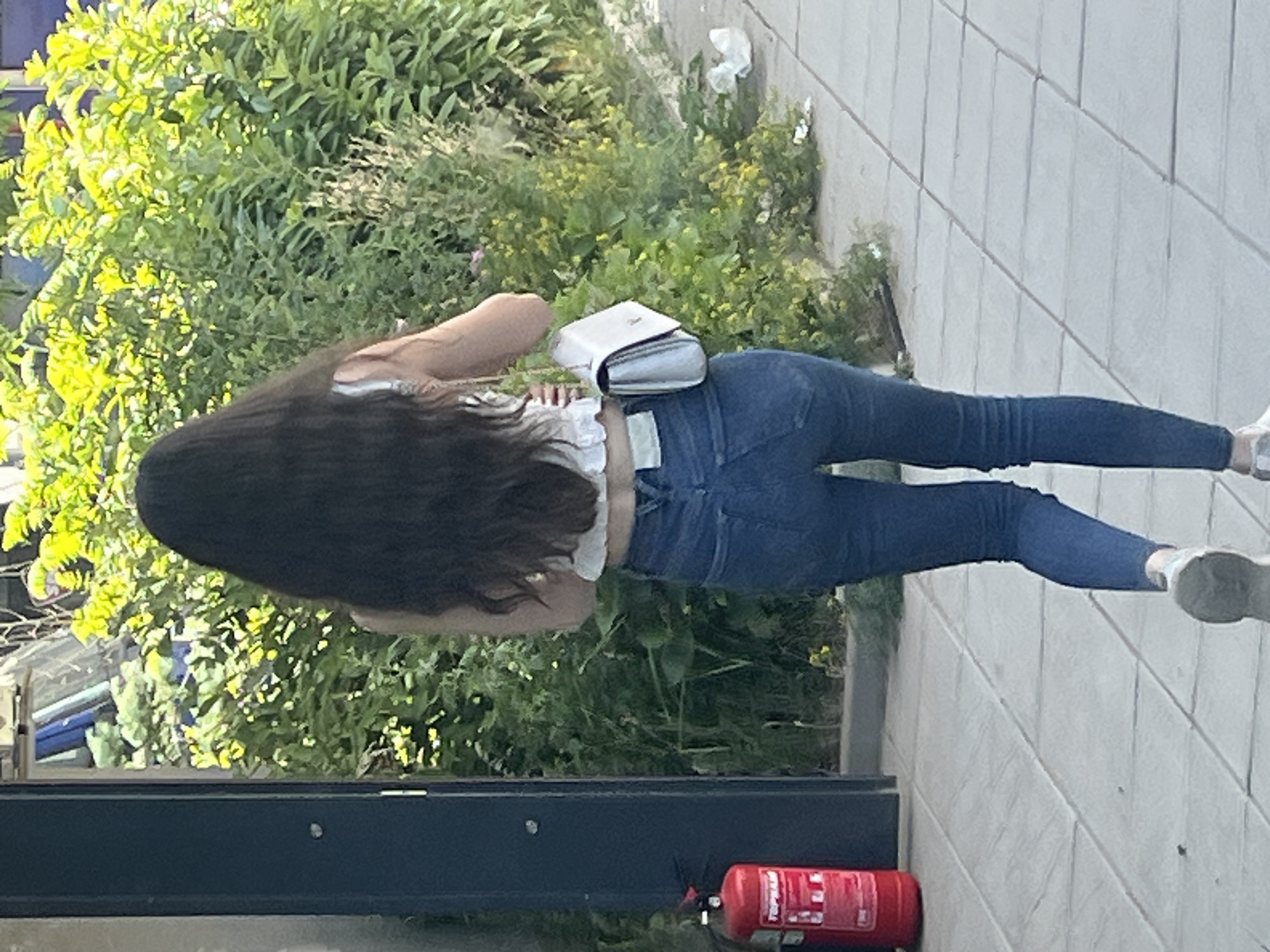 An outdoor full shot shows a young woman walking away from the camera, her dark, long hair obscuring her back. She is wearing a white ruffled crop top and blue denim jeans, and carrying a white purse over her right shoulder. The woman is walking on a paved sidewalk lined with green bushes and plants, with some trash visible on the ground. To the left of the frame, a red fire extinguisher is mounted on a dark blue pole. The background is out of focus, but shows greenery and a hint of a building. The lighting suggests it is daytime.