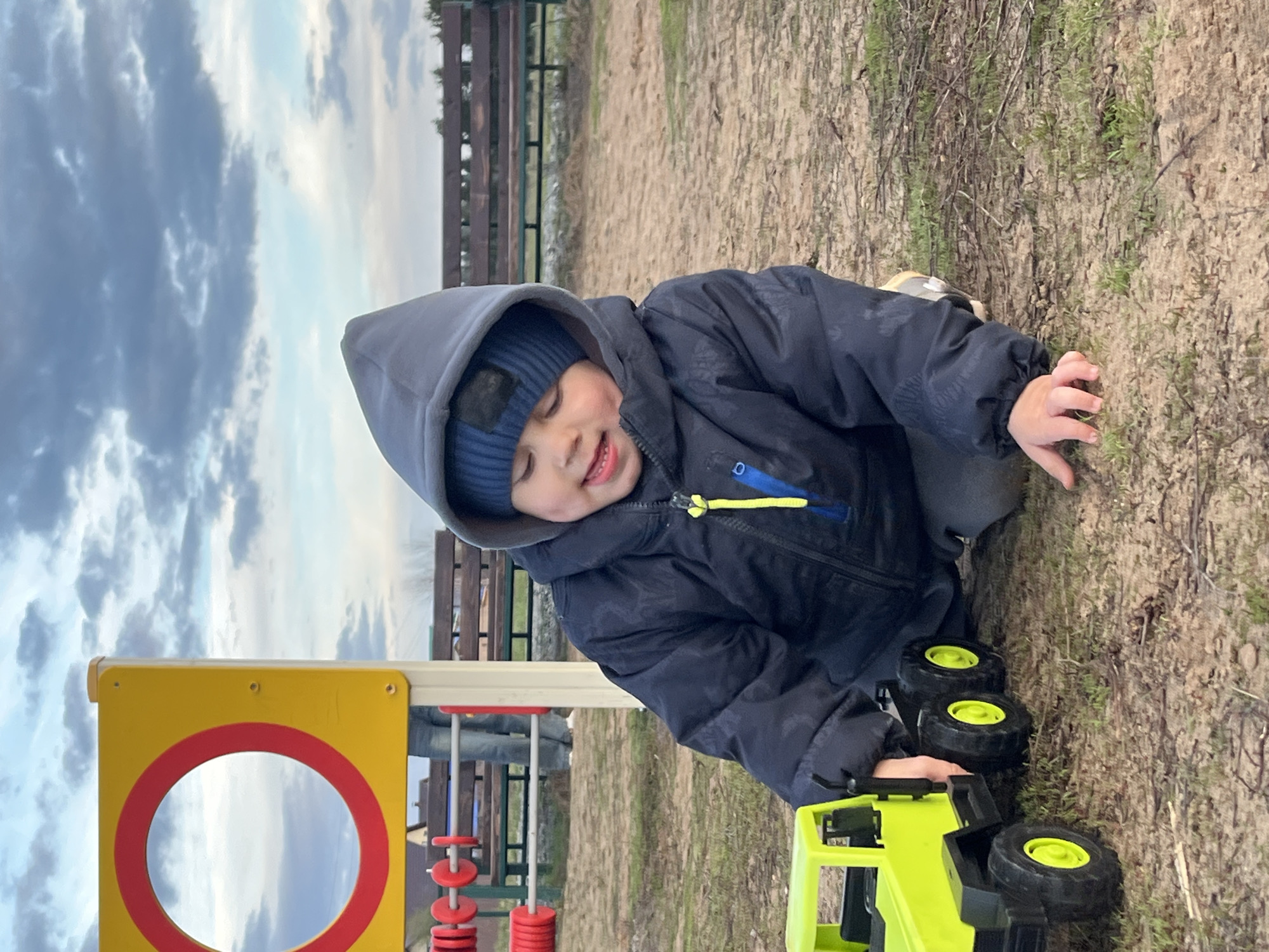 A young child in Фоки, Russia is playing with a lime green and black toy truck on a grassy and dirt area. The child is wearing a dark blue hooded jacket and a matching knit hat. The playground equipment is yellow and red and a weathered wooden fence is nearby. Overcast skies are visible.