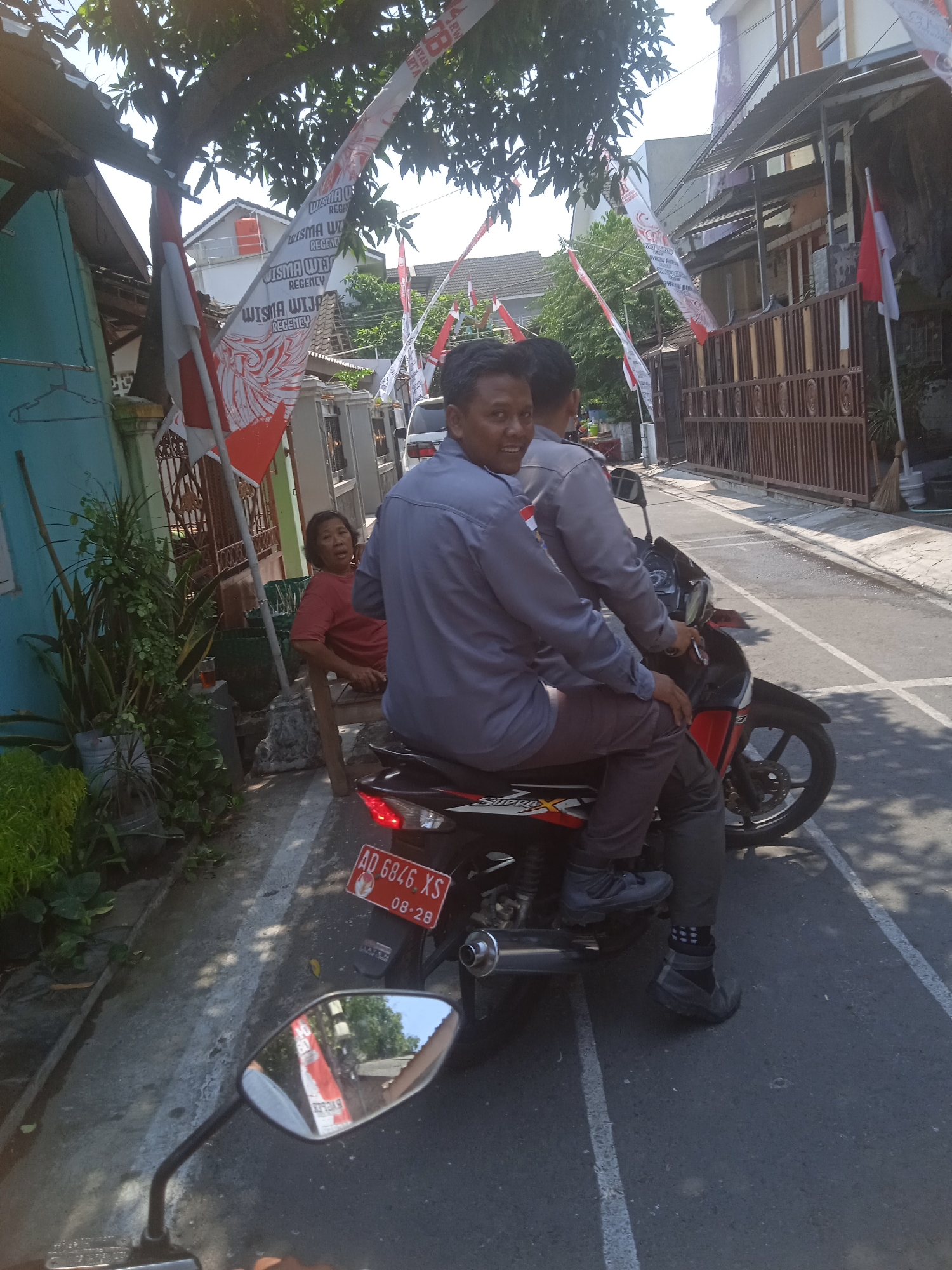 In Surakarta, Indonesia, two men are seated on a motorcycle on a paved street. The man in front is wearing a gray long-sleeved shirt and dark pants, and is looking towards the camera with a slight smile. The man behind him is wearing a gray long-sleeved shirt and dark pants, with one leg visible. The motorcycle is red and black, with a license plate reading "AD 6846 XS 08-28". A woman in a red shirt is sitting on a wooden chair to the left of the motorcycle. The street is lined with buildings and houses decorated with Indonesian flags and banners. There are also potted plants and trees visible. The scene appears to be a residential area on a sunny day.