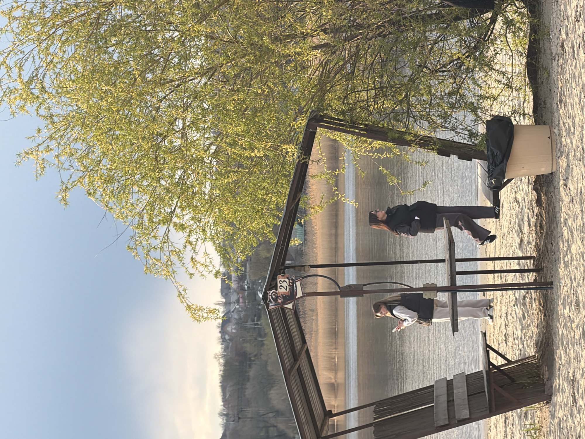 The scene is captured on a sunny day near a body of water, possibly a lake or a wide river, in Olginskaya, Russia. The image includes a man and a woman standing at the edge of the water, with a structure resembling a pier or a gazebo partially visible in the foreground. The gazebo has a sign with a raccoon illustration and the number "23". The terrain appears to be a sandy beach. The surrounding environment includes trees with budding leaves and a view of buildings on a distant hillside.