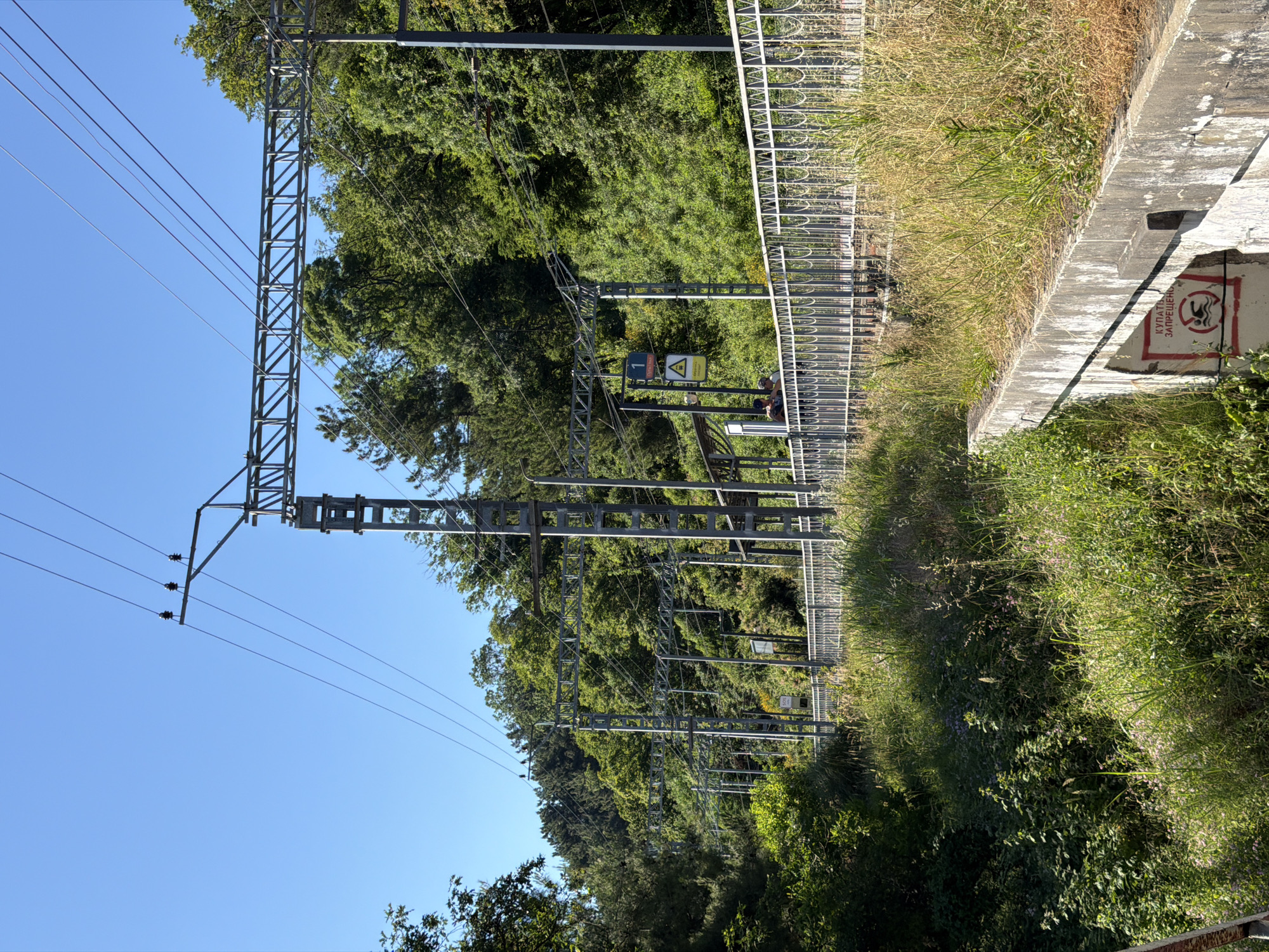The image shows a view of a railway track with overhead power lines, located in Sochi, Russia. A metal fence runs along the side of the track, separating it from a steep, vegetated embankment. Several people are visible standing near the fence, looking out over the tracks. The environment is lush and green, with trees and bushes growing on the hillsides. The sky is clear and blue. A sign on the embankment reads "КУПАТЬ ЗАПРЕЩЕНО", which translates to "SWIMMING PROHIBITED". The overall impression is a sunny day at a scenic, possibly remote, railway station.