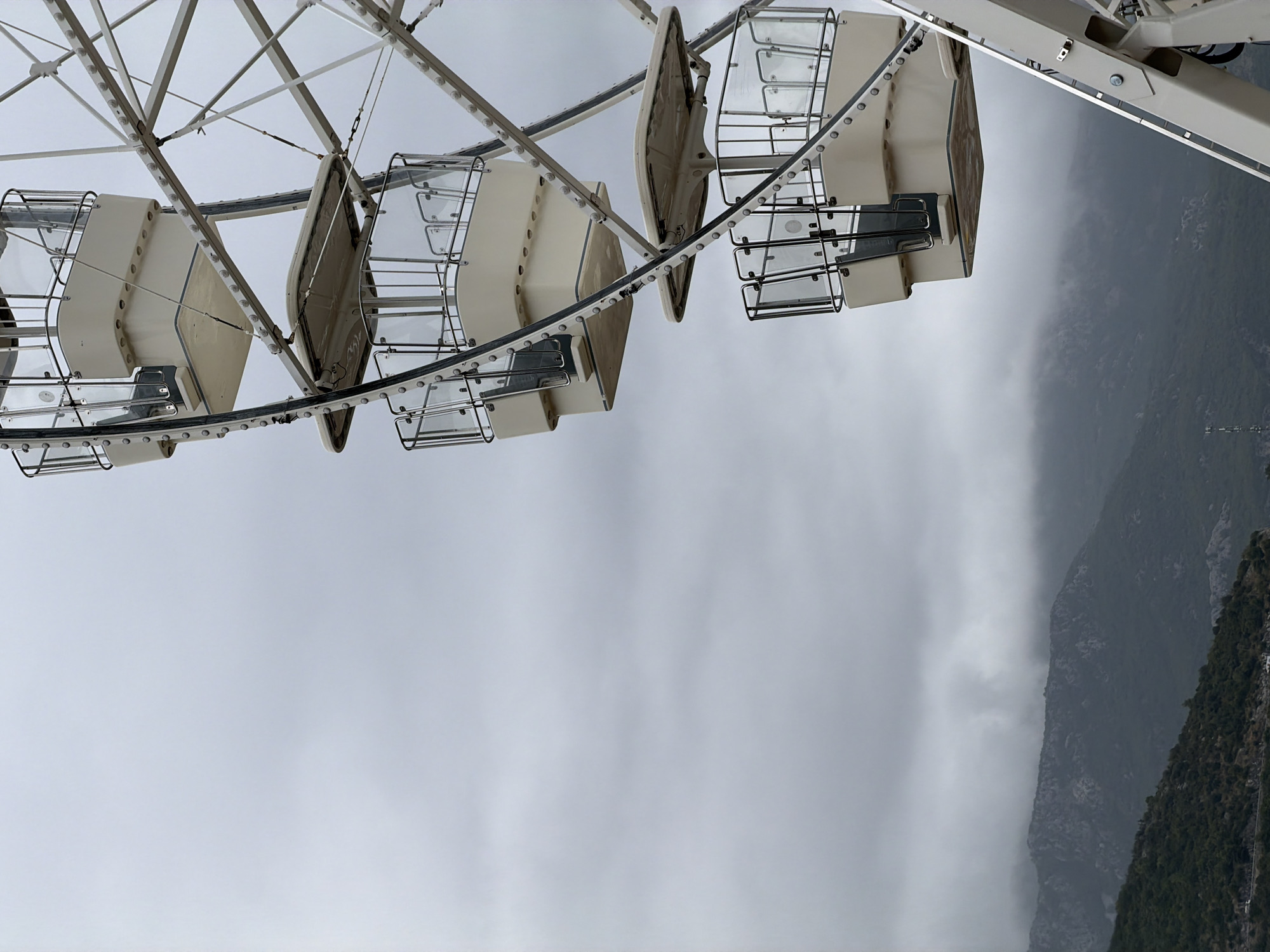 This is a close-up, vertical shot of a Ferris wheel against a cloudy sky, with mountains in the background. The Ferris wheel is white with beige gondolas, each equipped with glass panels and metal railings. Several gondolas are visible, stacked vertically. The landscape behind the Ferris wheel consists of dark, rocky mountains covered in green vegetation, shrouded in mist. The overall mood is overcast and slightly dramatic due to the weather and the imposing structures. The location is identified as Budva, Montenegro.