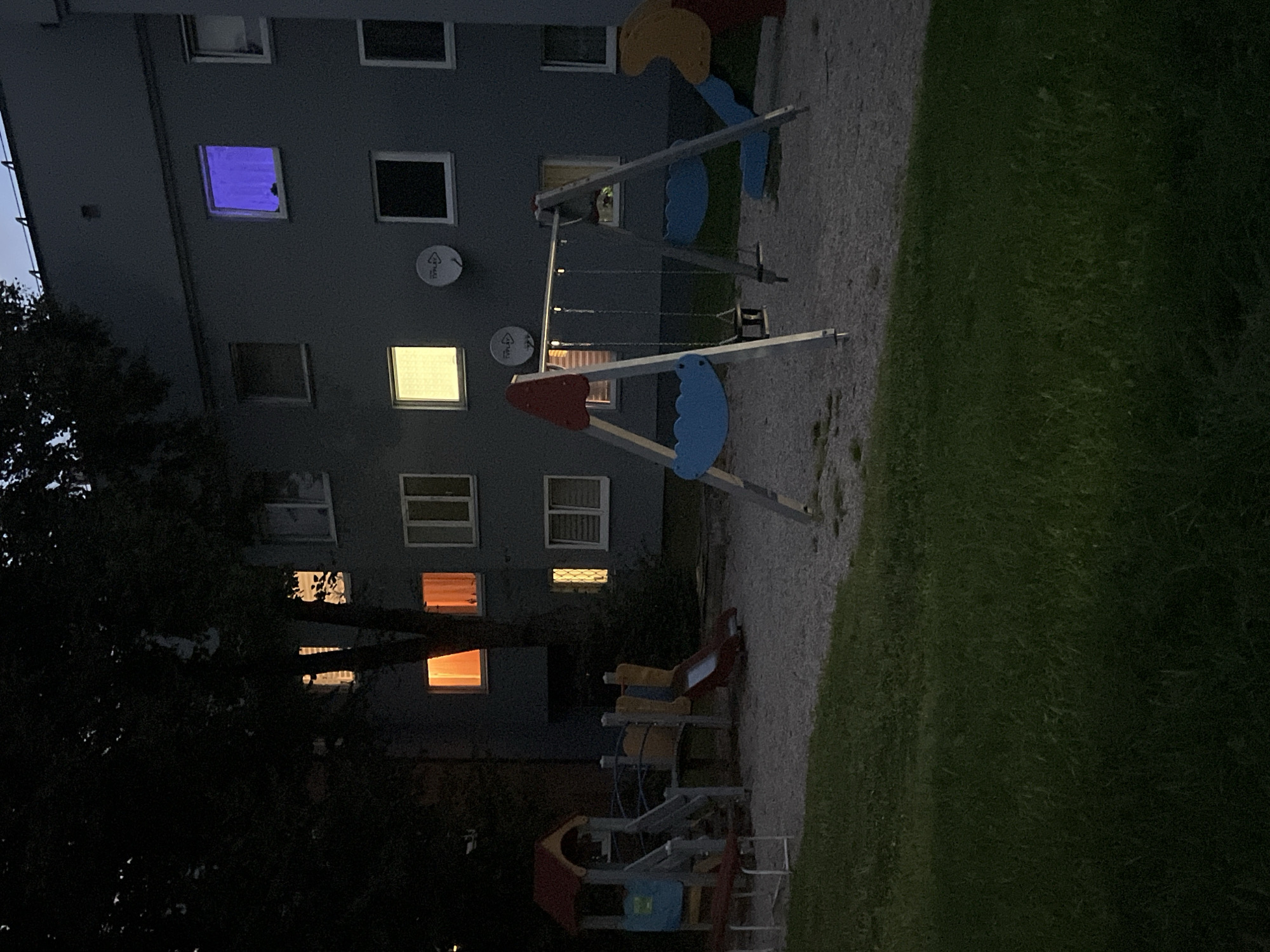 The image captures a dusk scene at a playground adjacent to a multi-story apartment building in Vilnius, Lithuania. The building, with its gray facade, features several windows, some illuminated from within. The upper left window on the second floor is bathed in a vibrant purple light, while other windows display yellow or orange glows. A satellite dish is visible on the building's exterior.
The playground itself is equipped with a swing set featuring two swings, a small multi-level play structure with a slide, and a red bench. The ground of the playground is covered with a light-colored granular material, likely for safety. To the left of the playground, a patch of green grass is visible, fading into darkness at the bottom of the frame. A large tree with dense foliage partially obscures the building and playground. The overall atmosphere is quiet and still, with the lighting suggesting twilight or early evening. No people are present in the scene.