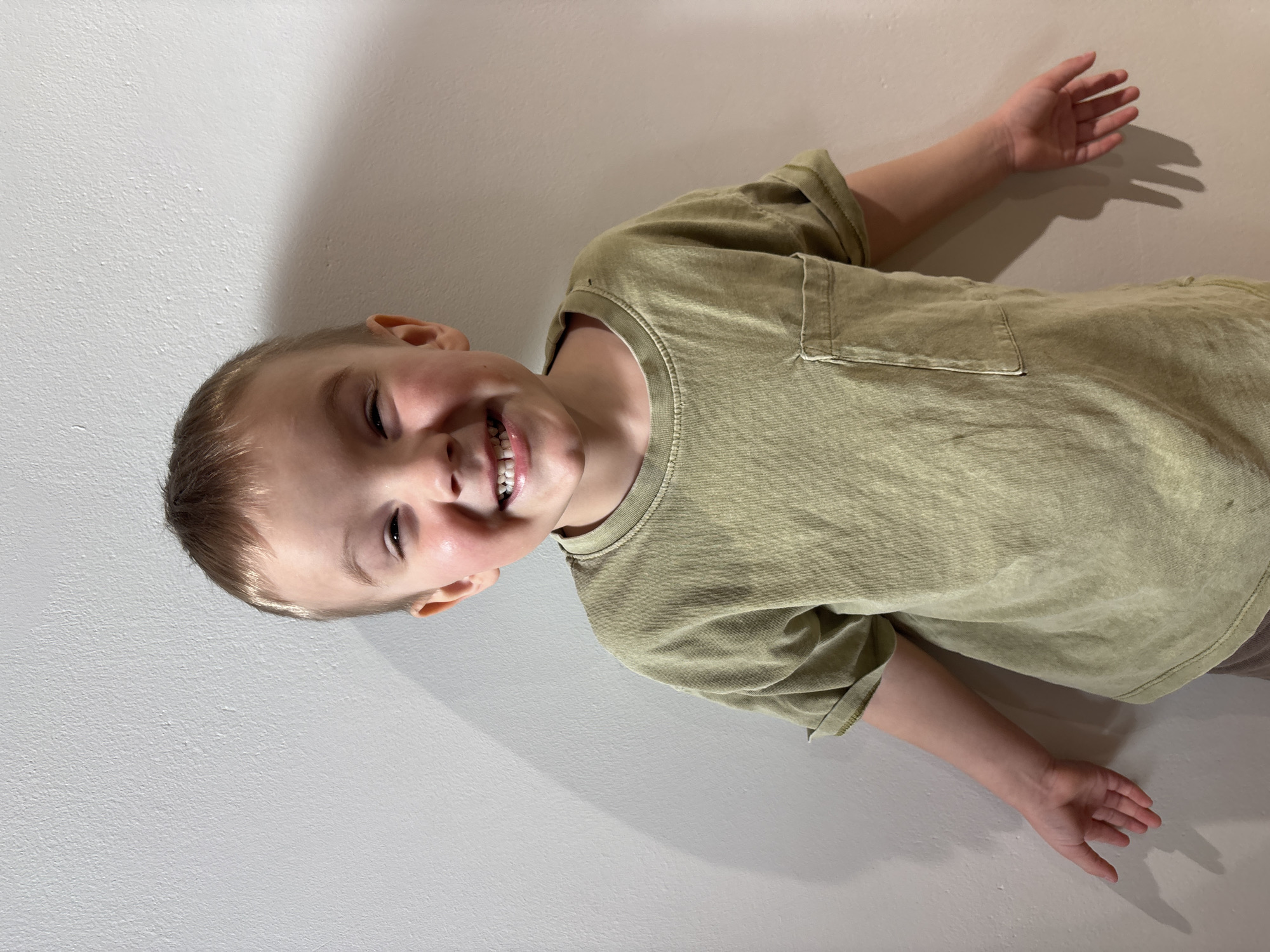 A young boy with short blonde hair and a bright smile is lying down with his arms outstretched against a pale wall. He is wearing a light green t-shirt with short sleeves and a chest pocket. Shadows are cast on the wall behind him, indicating a light source from above. There is no visible evidence to suggest the location is Chaikovsky, Russia.