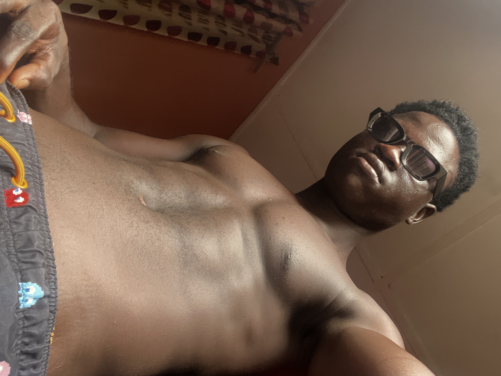 An individual with dark skin and short, curly hair is shown wearing dark sunglasses. The person appears to be shirtless, wearing shorts with a dark background and colorful patterns including what may be Pac-Man characters. The background includes an orange-painted wall and a patterned bedspread. The person's location is specified as the city of Mosafejaw, Nigeria, but there are no visual elements in the image to confirm this location.