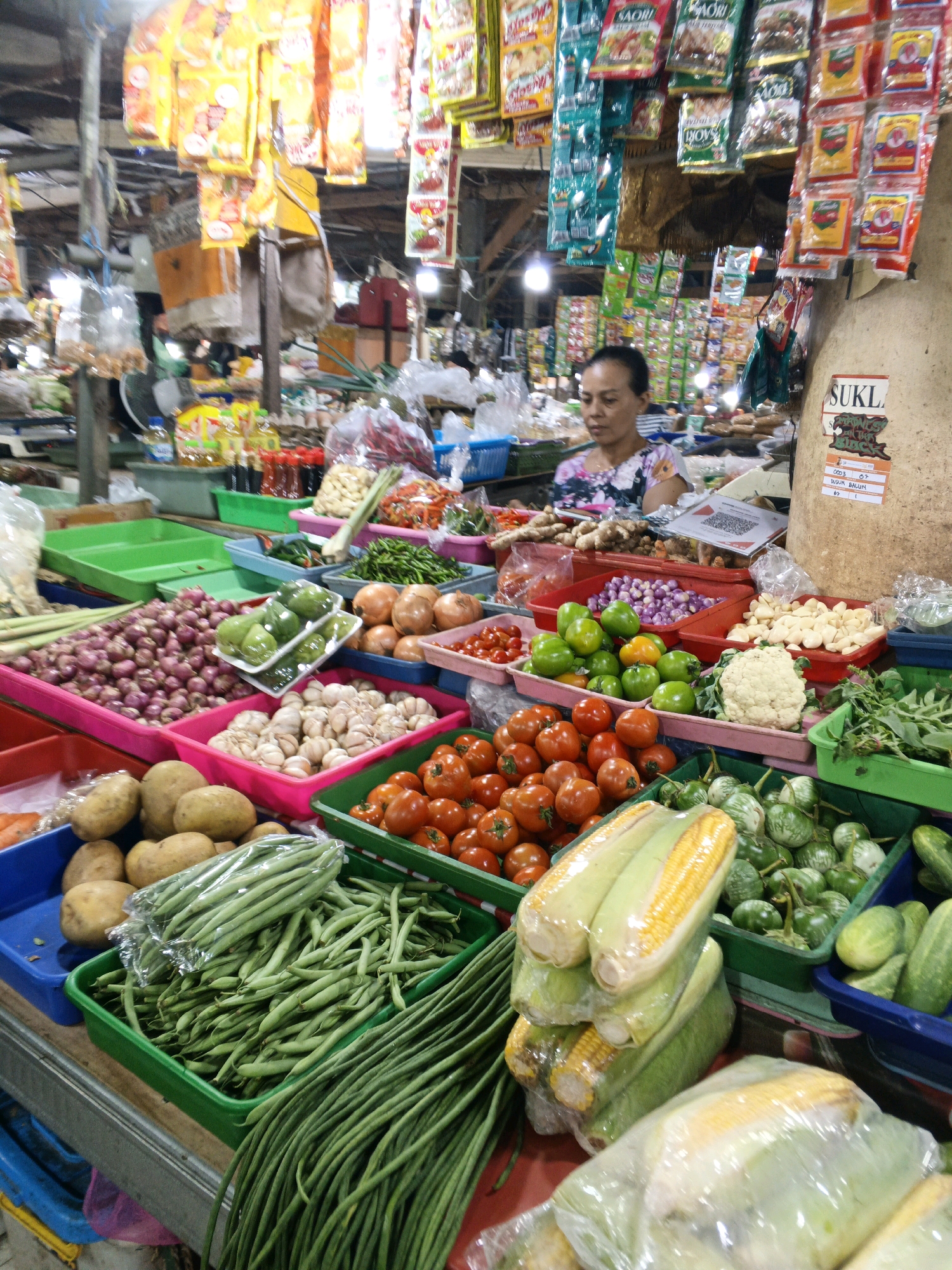 This image depicts a bustling market stall in Denpasar, Indonesia, likely a traditional wet market. The stall is laden with a vibrant assortment of fresh produce, meticulously arranged in various plastic containers and baskets. In the foreground, neatly stacked bunches of green beans and long beans are prominent. Behind them, rows of ripe tomatoes, green chilies, and potatoes are visible, alongside heads of cauliflower and a variety of onions and garlic. There are also displays of corn on the cob, some wrapped in plastic.
In the mid-ground, a woman with dark hair pulled back is seen behind the counter, appearing to be attending to customers or organizing the produce. She is wearing a floral patterned shirt. The market environment is characterized by overhead lighting, visible through a semi-transparent ceiling structure, and an abundance of packaged goods hanging in the background, including brightly colored food products and spices. The overall impression is one of abundance and lively commerce, typical of Indonesian markets.