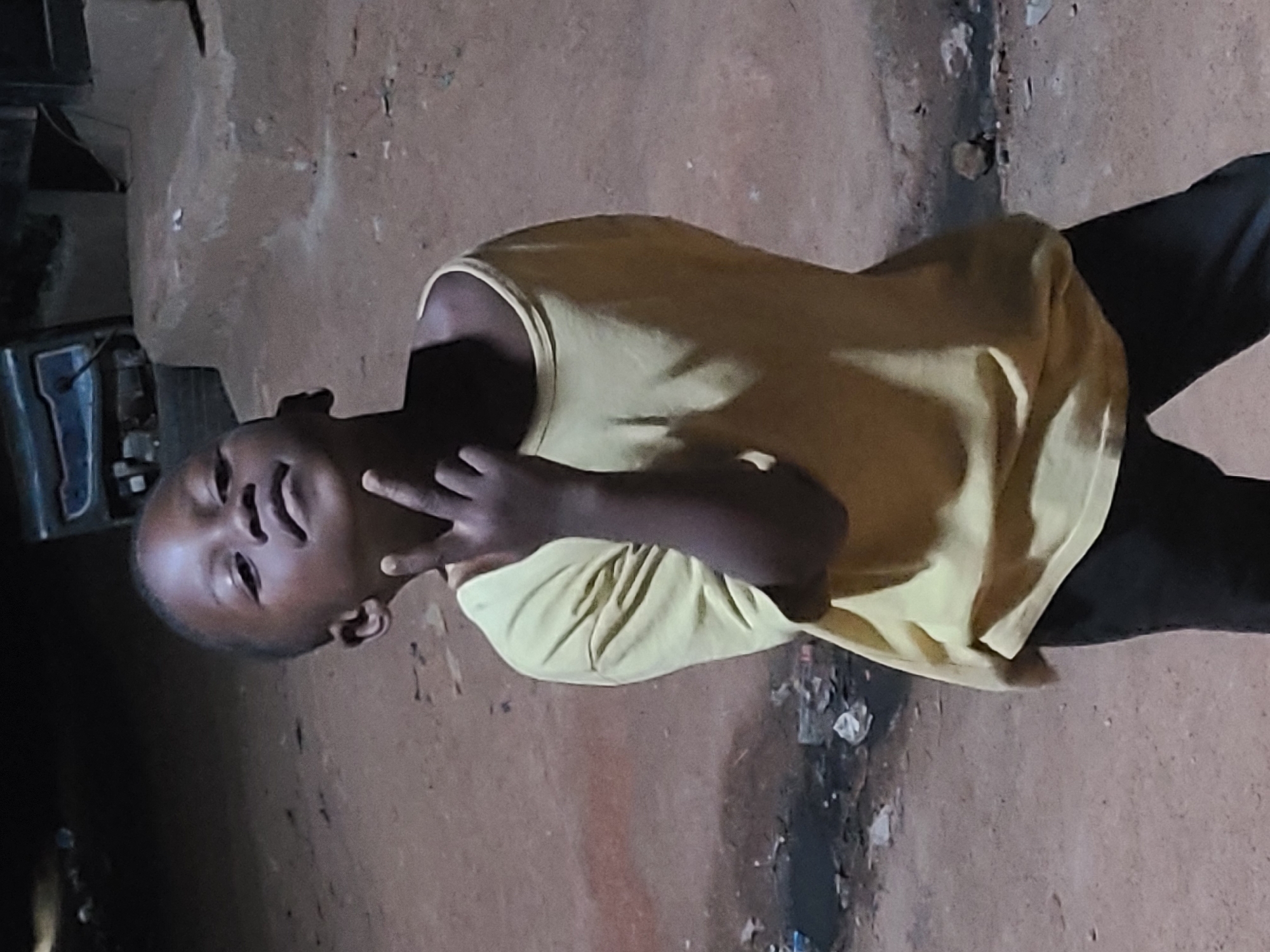 A young person wearing a yellow shirt and dark pants stands on what appears to be a dirt ground outside in a dimly lit environment. The individual is gesturing with their fingers in a "peace" or "victory" sign. Debris is scattered on the ground to the right of the person. In the background on the left, there's a glimpse of some unidentified structure or object, possibly a building or a piece of equipment. The general environment suggests the location could be a residential or semi-urban area. I can't confirm the location as Jos, Nigeria, without additional information.