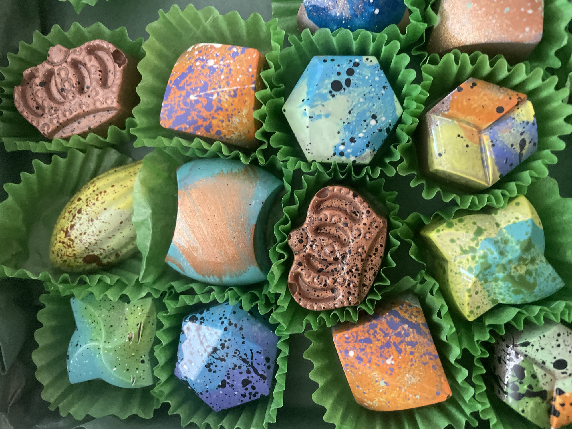 The image shows a box of twelve artisanal chocolates nestled in green paper cups. The chocolates are various shapes, including a crown, hexagon, cube, and rounded forms. They are decorated with colorful patterns and splatters, featuring hues of orange, blue, purple, yellow, green, and brown. The lighting is bright and the focus is sharp, showcasing the intricate details of the chocolates. No people, city locations, actions or interactions are visible in the photograph. The environment consists solely of the chocolates and their packaging.