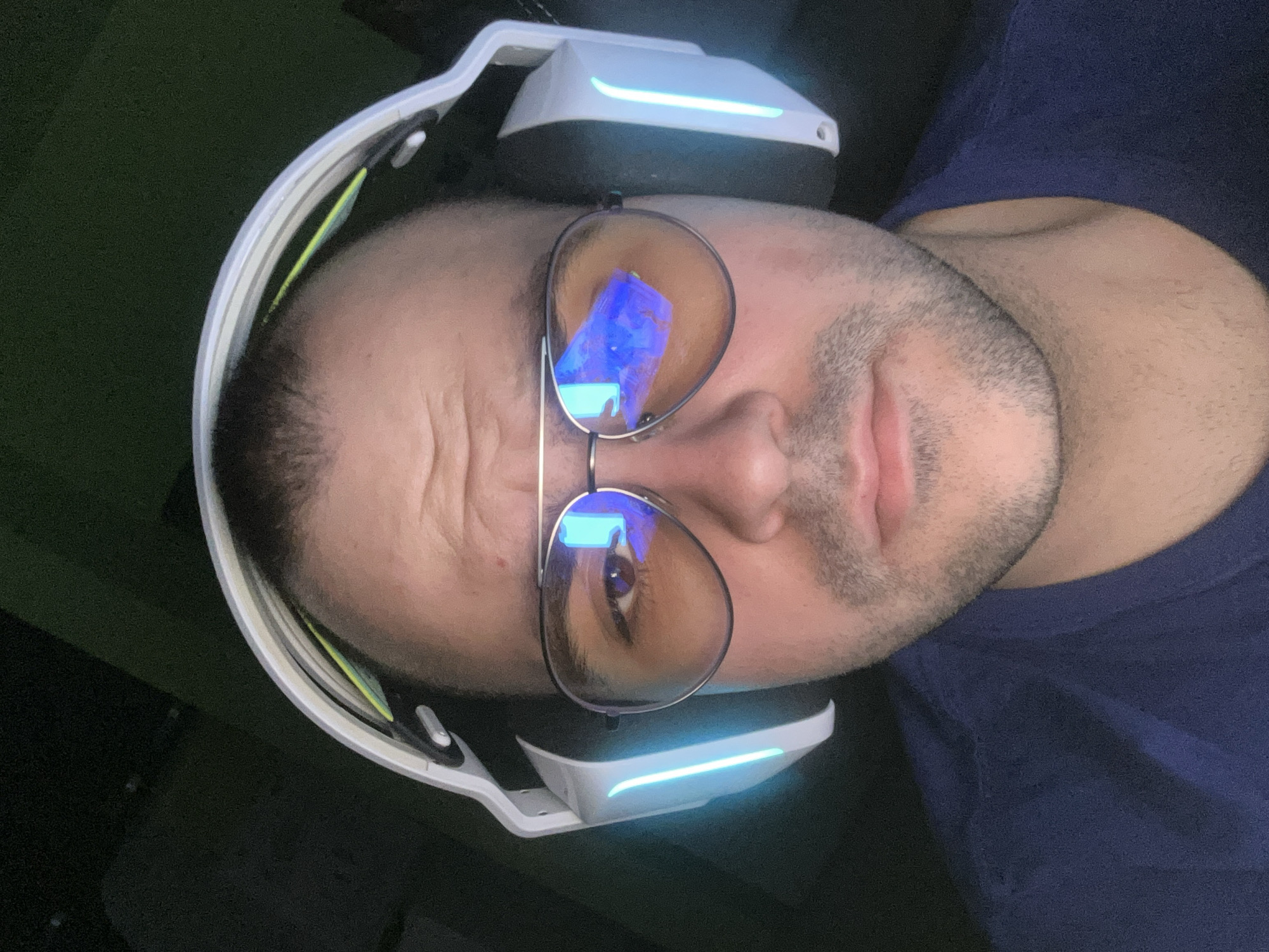 A close-up, rotated image shows a man's face. He is wearing aviator glasses with reflective lenses. He is also wearing white headphones with blue lights on the sides. The man has short, dark hair and a light complexion. He is wearing a dark blue t-shirt. The background is dark and indistinct. There is no information available to determine if the image was taken in Plovdiv, Bulgaria.