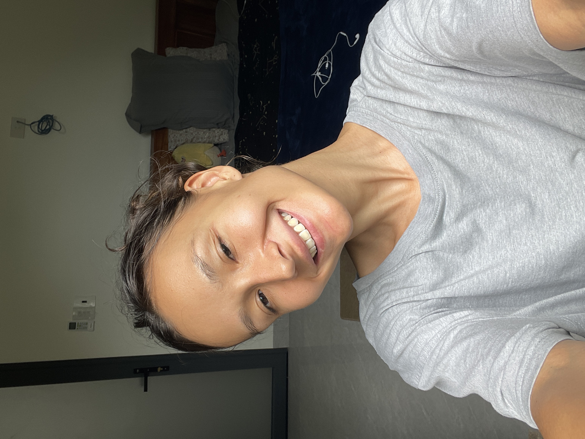 An adult individual, likely female, takes a close-up selfie indoors, smiling broadly at the camera. Her dark hair is pulled back, and her face shows a natural, happy expression, highlighting her white teeth. She wears a simple, light grey t-shirt.
The background reveals a casual interior space, possibly a bedroom. To the viewer's left, a light-colored wall features a white electrical outlet with a coiled blue cable hanging from it. Further right, a bed is partially visible, covered with a dark blue blanket or duvet that has a faint white pattern. A grey pillow rests on the bed, and a small, light yellow object is tucked near the headboard. White wired earphones are casually placed on the dark blue bedding. At the bottom of the frame, a dark horizontal bar, possibly part of furniture or a door frame, is present. Another white electrical component is mounted on the wall behind the person's left eye.
The lighting suggests an indoor setting, likely during the day with diffused light, or in an artificially lit environment. No specific weather conditions are discernible. While the image itself lacks explicit location cues, the provided context indicates the location is Mũi Né, Vietnam. The person's joyful smile conveys a sense of contentment and happiness. There is no visible text in the image.