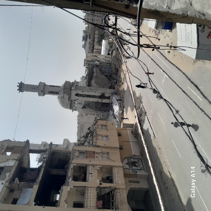 The image displays a severely damaged urban landscape in Khan Yunis, Palestinian Territory, under clear daylight conditions. The central focus is the Great Mosque of Khan Yunis in the mid-ground, which is extensively damaged; its dome and minaret remain partially standing, but significant sections of its walls are collapsed, exposing interior structures. To the left, a multi-story building, appearing residential or commercial, is largely destroyed, with multiple floors breached, walls crumbled, and rebar visible. Debris is scattered around the bases of both structures.
An asphalt road runs horizontally across the foreground and mid-ground, featuring faded white lane markings. Scattered small piles of rubble are visible along the road's edges. Overhead, numerous black electrical or communication wires crisscross the frame, appearing haphazardly strung.
Several individuals are present: on the left side of the road in the foreground, one person walks away from the viewer, casting a long shadow. Further down the road, an adult in dark clothing and a smaller child in red clothing walk towards the mosque. In the area surrounding the mosque, near white tarp-like structures that may be temporary shelters or collection points, several indistinct figures are visible amidst the debris. The image is captured from an elevated perspective, looking down onto the street and across the scene. A "Galaxy A14" watermark is visible in the lower left corner.